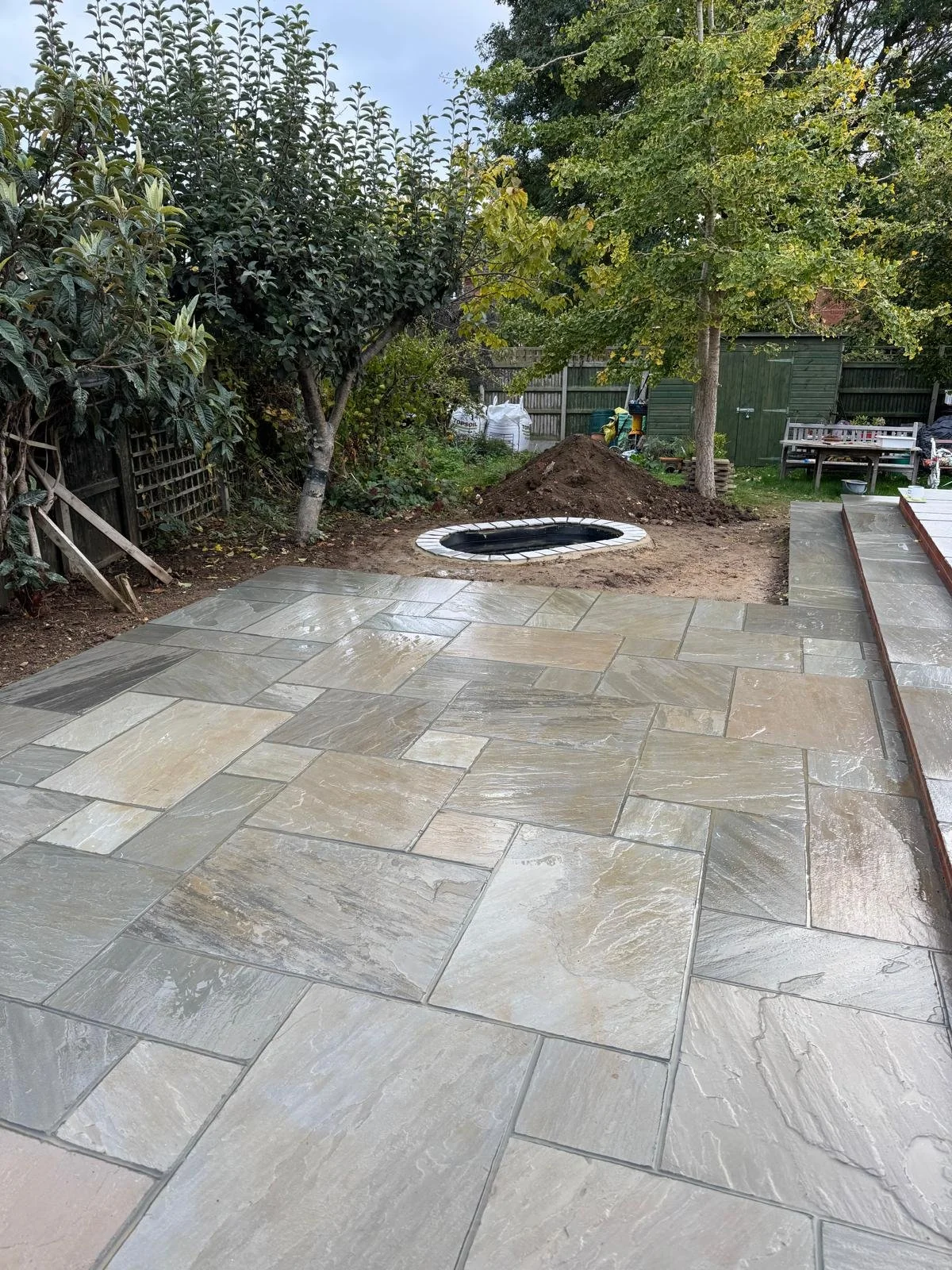 Backyard with a new paved stone patio, a small dug hole surrounded by white bricks, a raised garden bed on the right, trees, and a garden shed in the background.