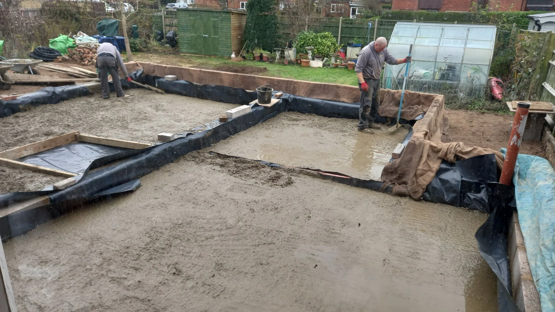 Two people are working in a backyard, laying a concrete foundation, with one spreading wet concrete and the other preparing the area with tools and boards.