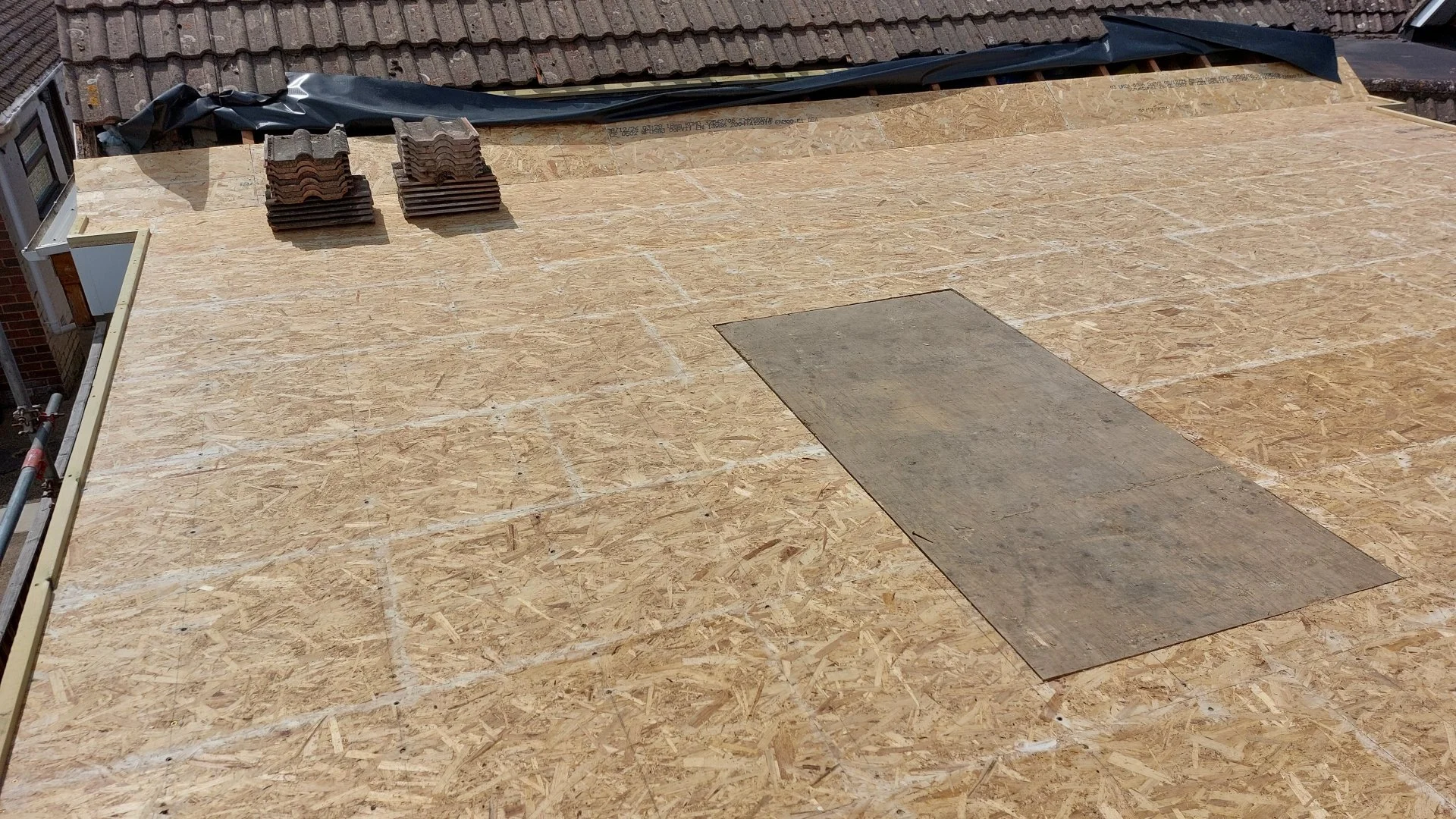 Flat roof covered with oriented strand board (OSB) sheathing, with stacks of roofing tiles and a rolled roofing membrane on top, and a rectangular plywood patch.