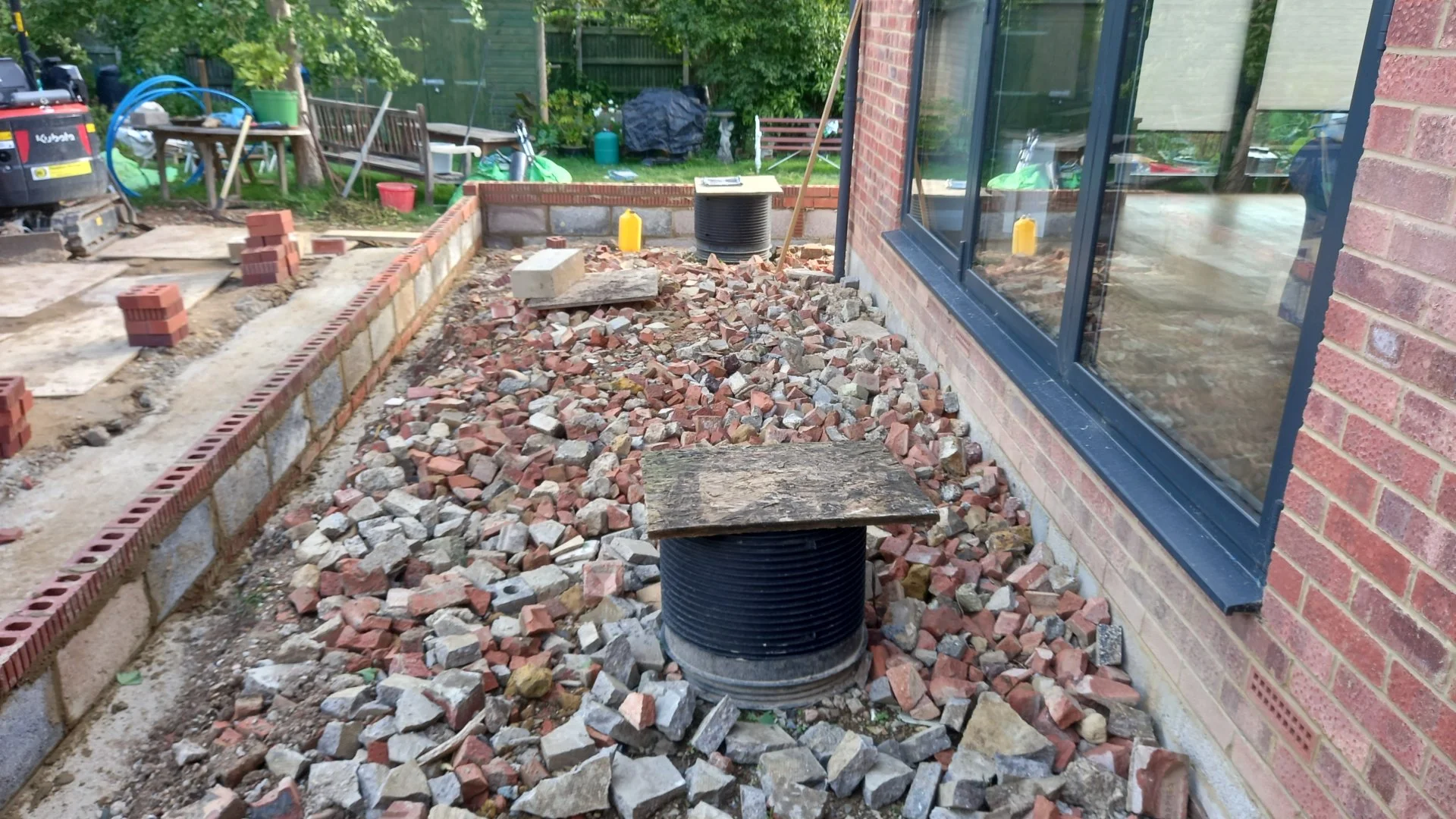 Construction site of a brick walkway outside a house. Partially built brick wall with sand and pile of small bricks nearby. Large window with black frame and brick wall of the house visible.
