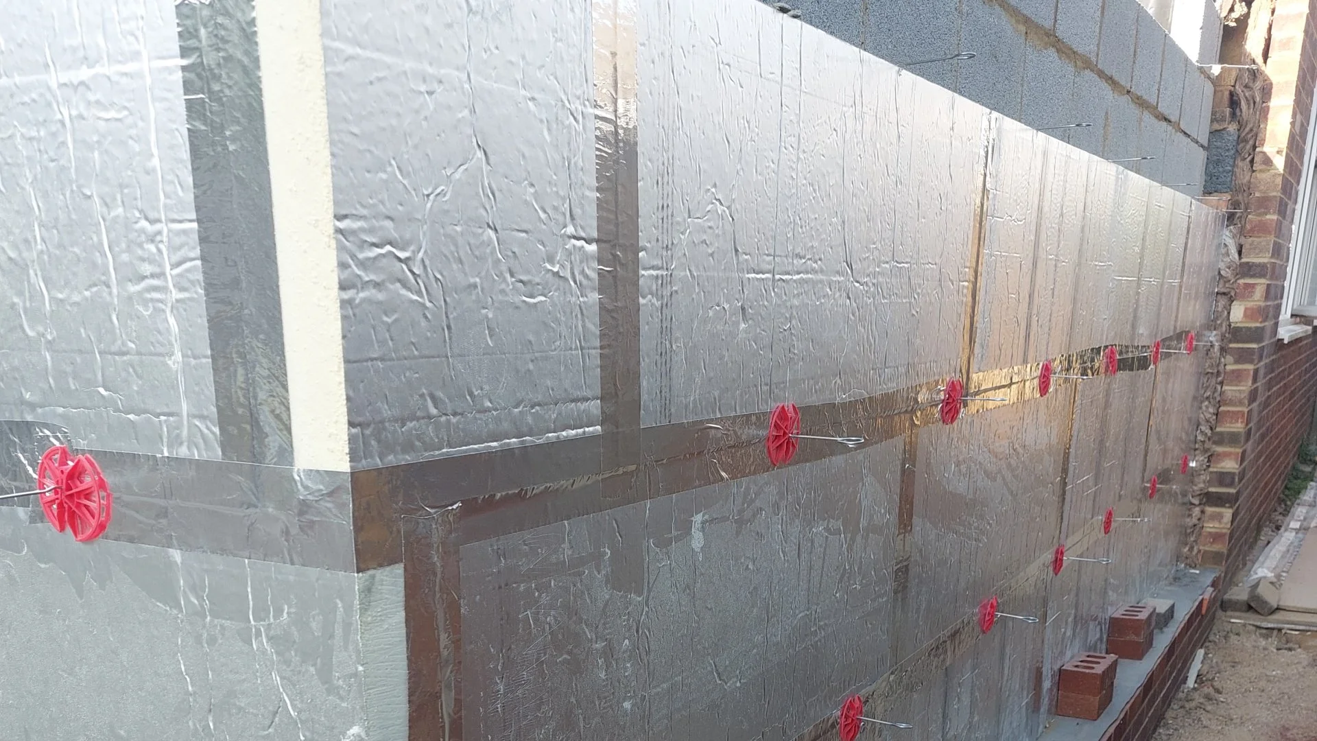 Installing wall insulation with shiny foil facing in a construction site, using red plastic spacers to maintain gaps between insulation sheets.