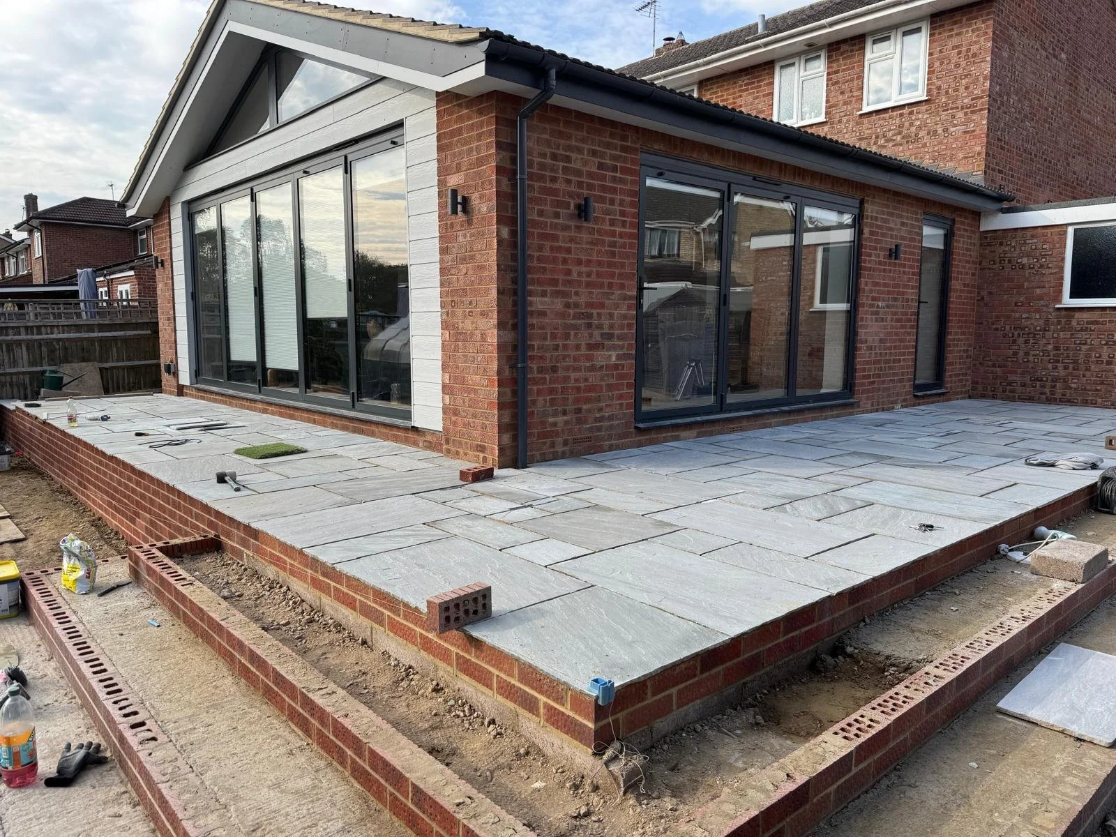A house with a brick exterior and large sliding glass doors, surrounded by an ongoing patio construction with paving stones and tools.