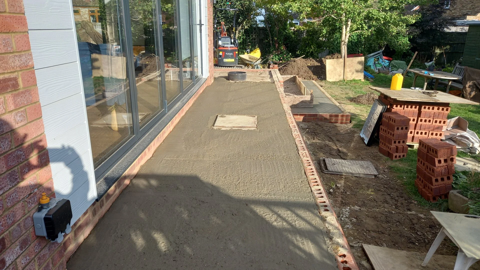 A backyard patio under construction with a cleared concrete slab, bricks along the edge, and construction tools and materials such as stacked bricks, a yellow container, and a table with tools. There's a large window on the house and landscaping with