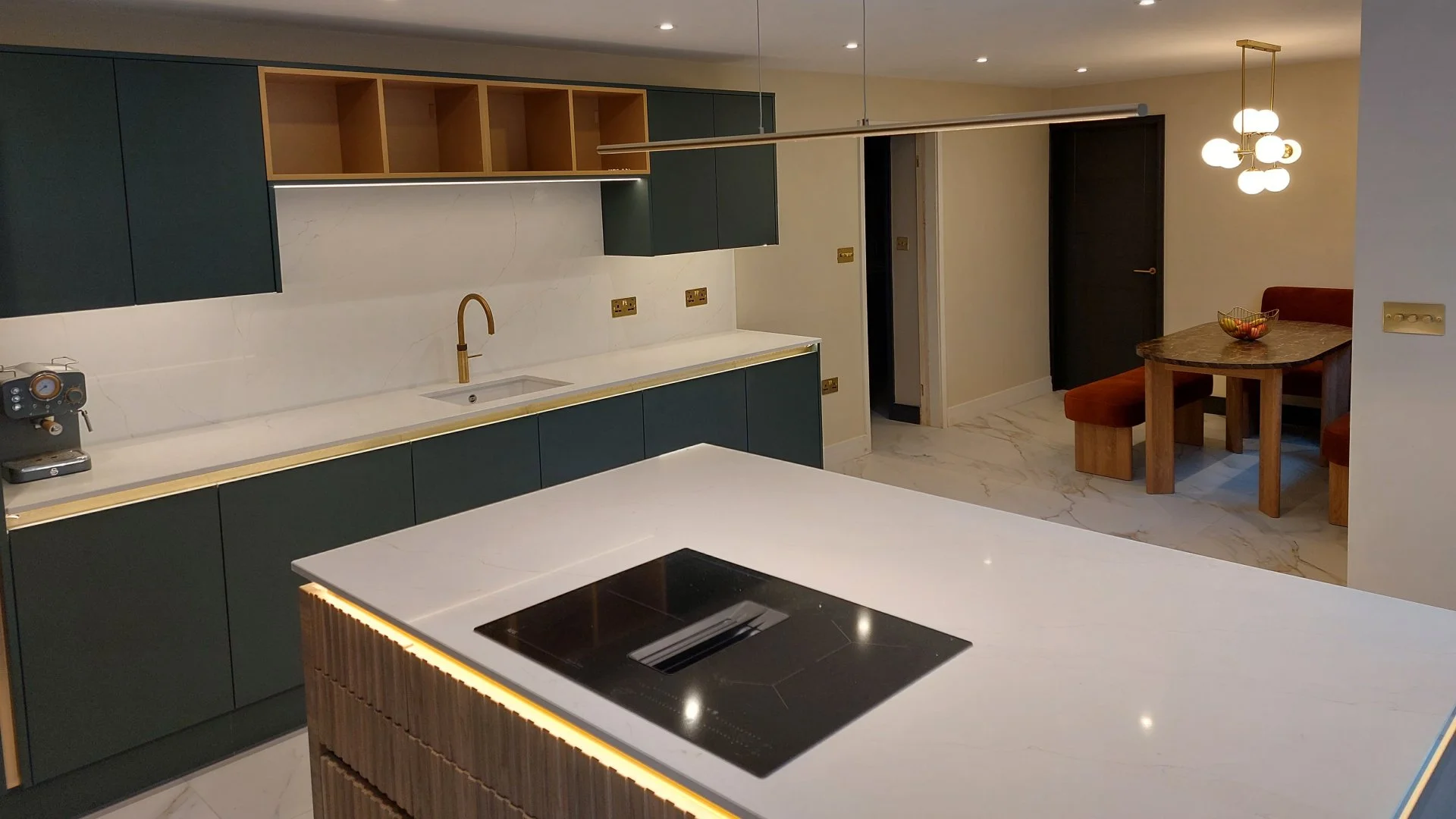 Modern kitchen with dark green cabinets, a white marble backsplash, a gold faucet, and a white island with a black cooktop. In the background, there is a dining area with a wooden table, red cushioned chairs, and a modern chandelier.
