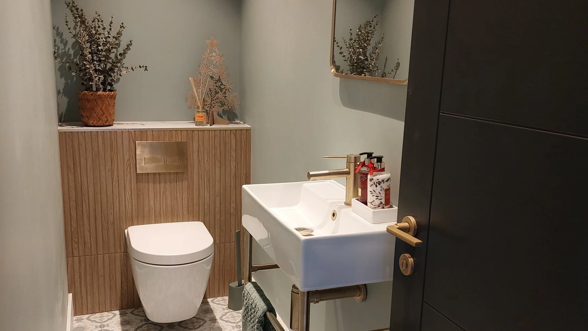 Small modern bathroom with a wall-mounted toilet, a small white sink with gold-colored fixtures, a black cabinet, and decorative items including a potted plant and a miniature Christmas tree on a wooden cabinet, with a mirror on the wall.