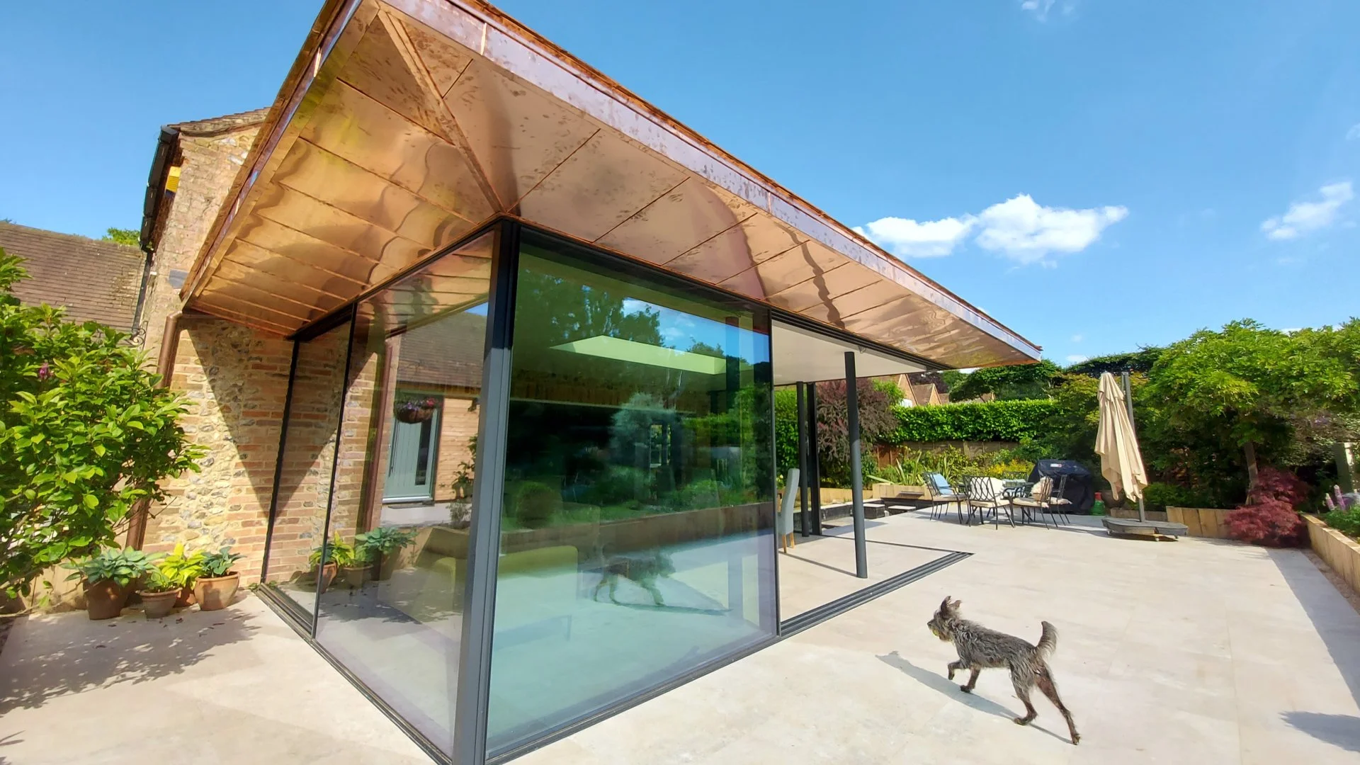 Modern house with large glass windows and a copper roof overhang, situated in a backyard with outdoor furniture, umbrellas, potted plants, and a dog walking on a concrete patio under a clear blue sky.