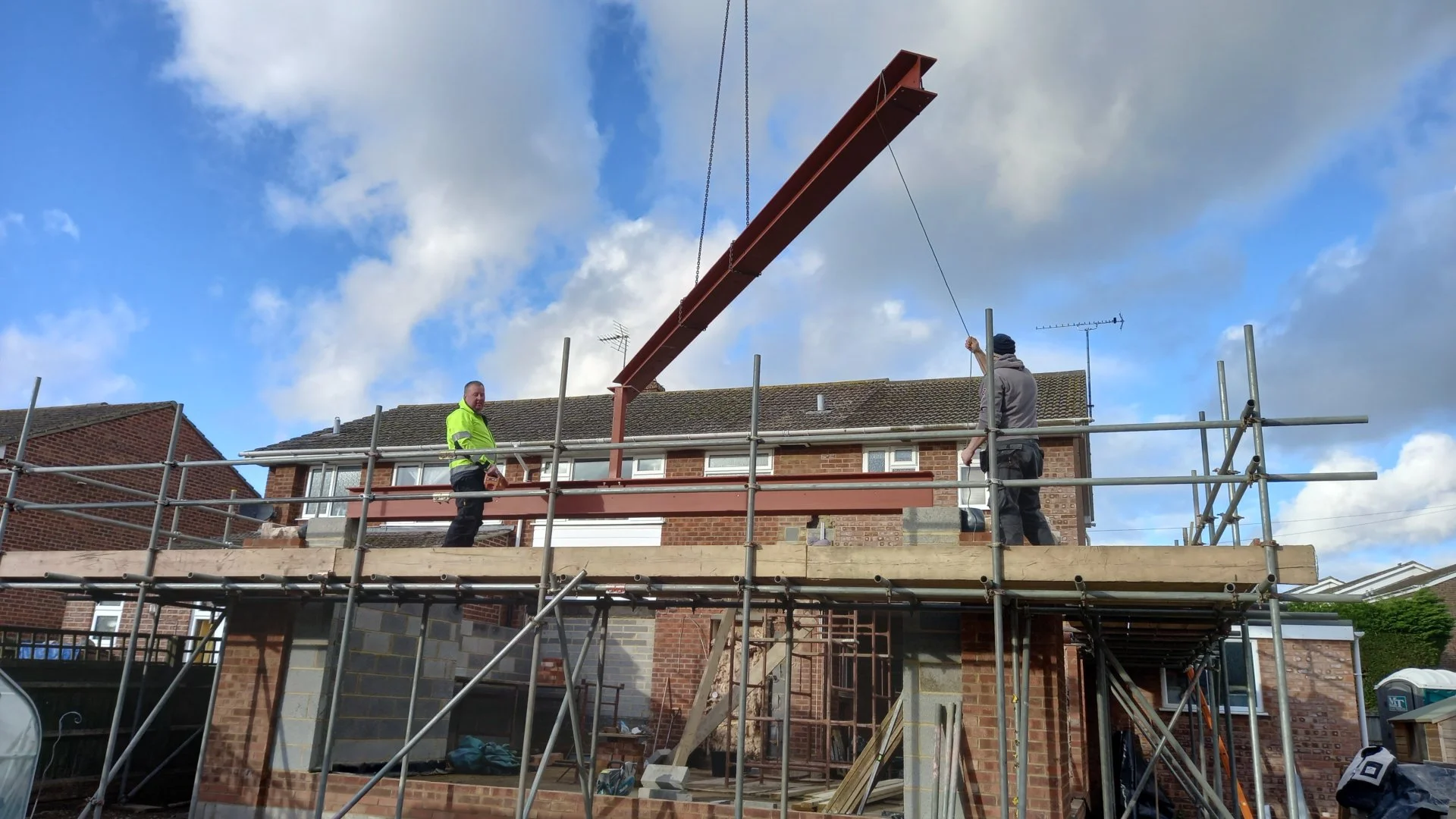 Construction workers installing a steel beam on a building's second floor. The scene shows scaffolding, a brick house, and two workers handling the beam under a partly cloudy sky.