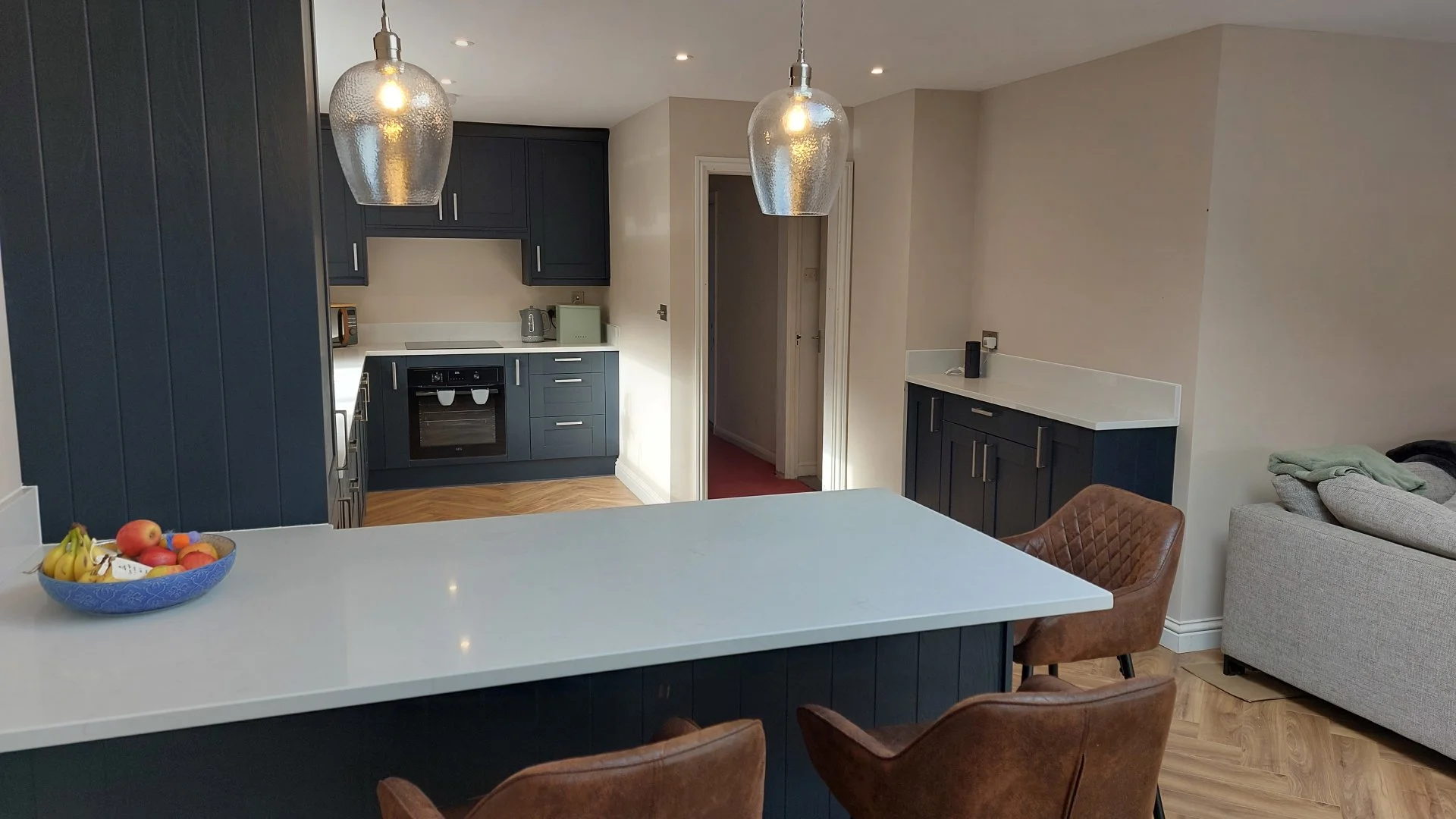 Interior view of a modern kitchen with navy blue cabinets, white countertops, and two hanging pendant lights. There is a fruit bowl on the counter, a gray sofa, and a living area visible in the background.