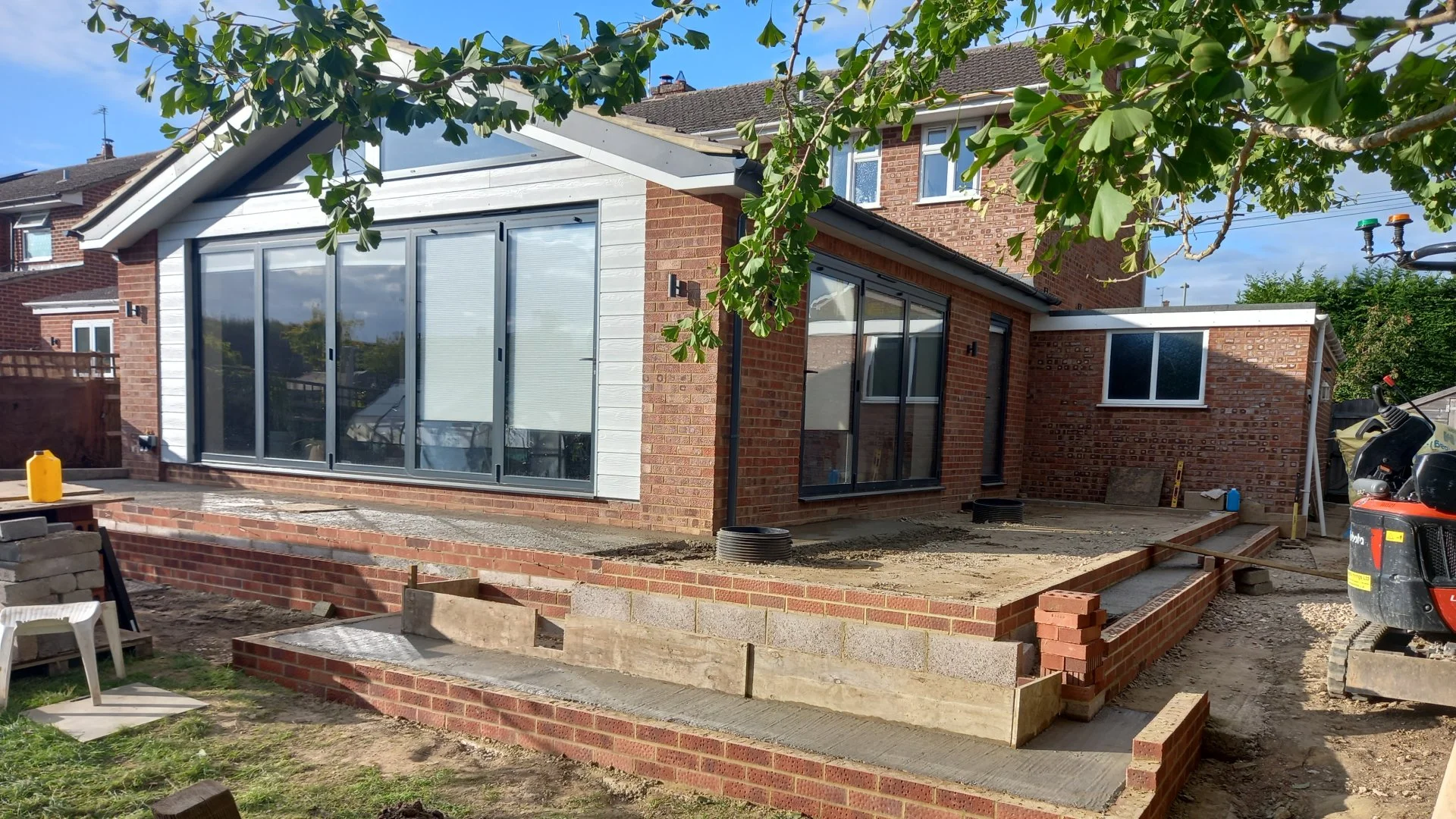 A house under construction with brick and white siding walls, large glass sliding doors, and construction tools and materials on the ground, including a small excavator and a plastic chair, with a tree overhead and a neighboring brick house in the ba