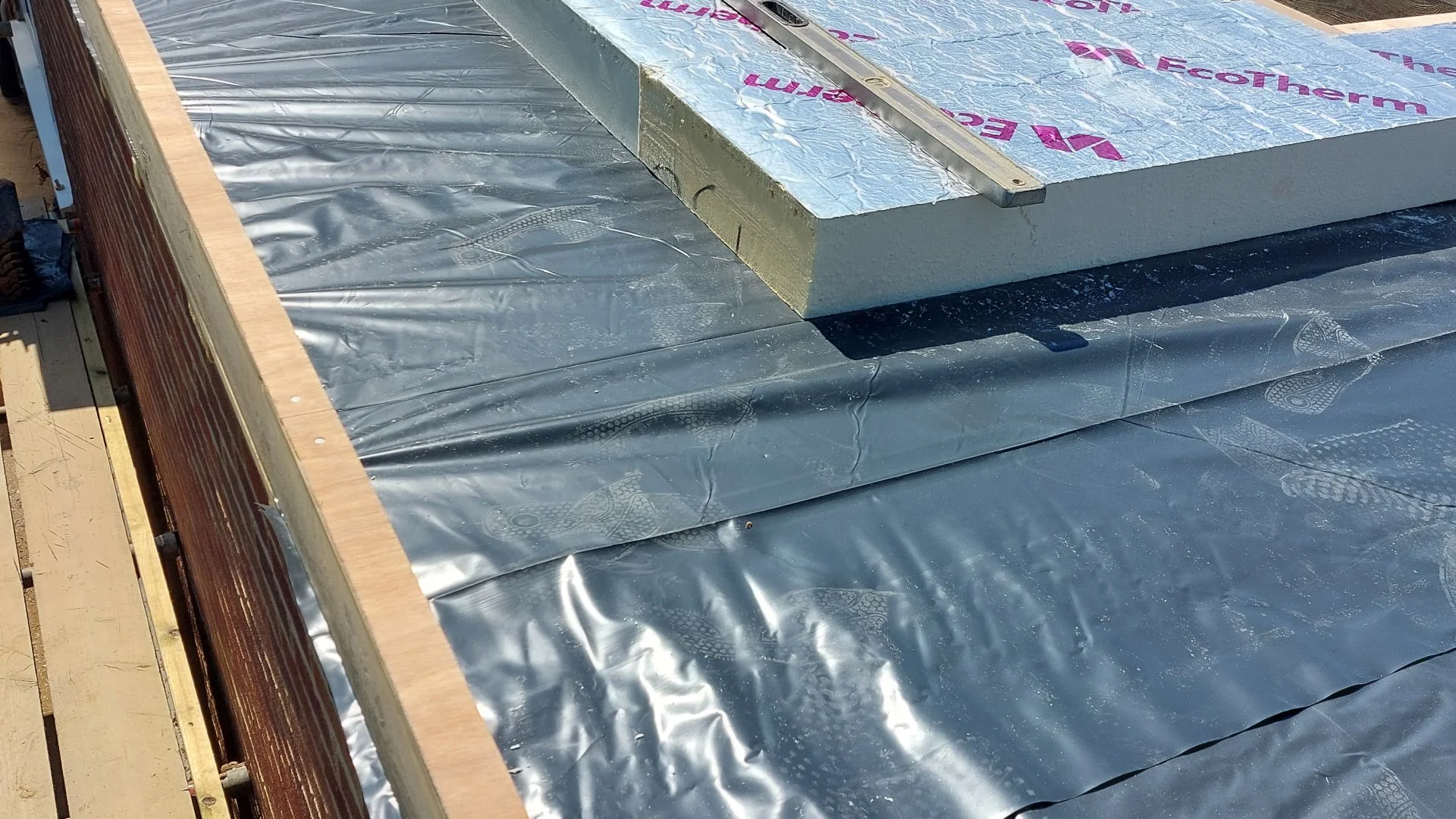 Construction site with roofing materials, including a wooden edge, metal flashing, and insulation foam with pink branding.