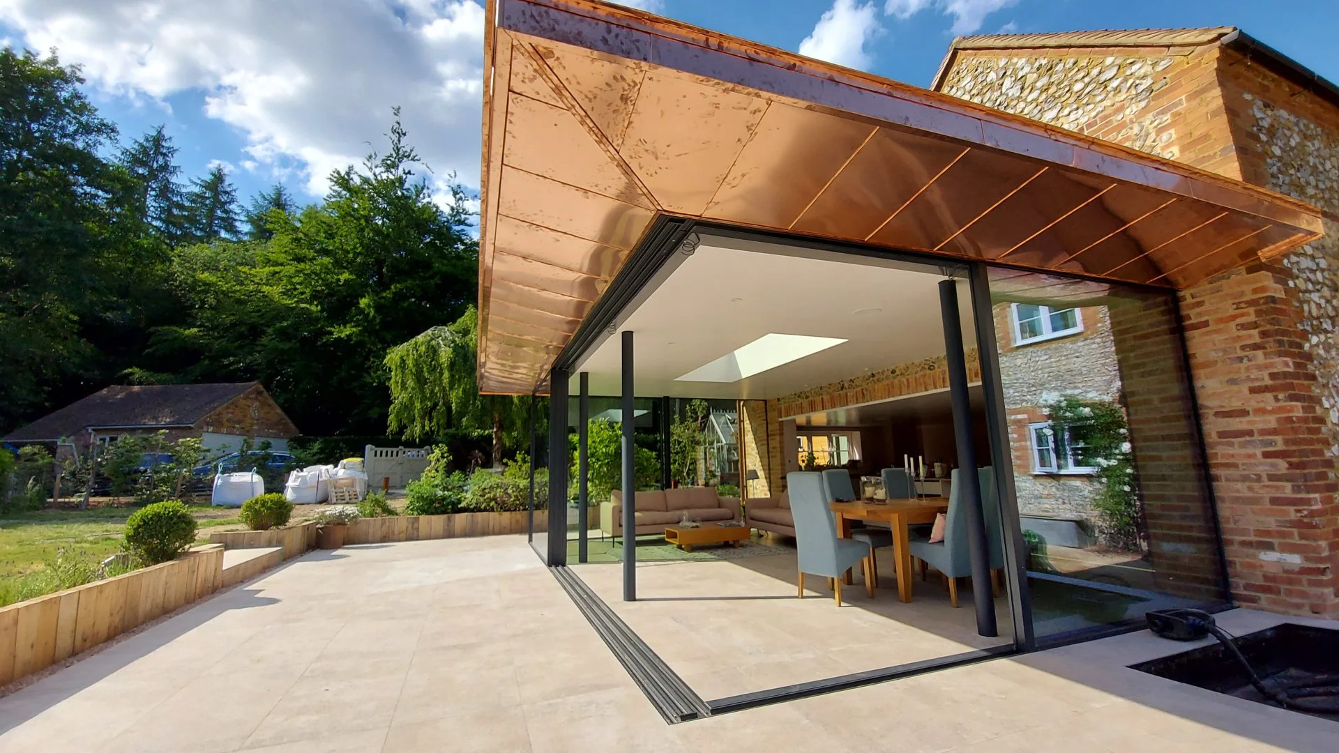 Modern house with large glass doors opening to a patio, outdoor furniture, and a garden with trees and shrubs, under a sky with clouds.