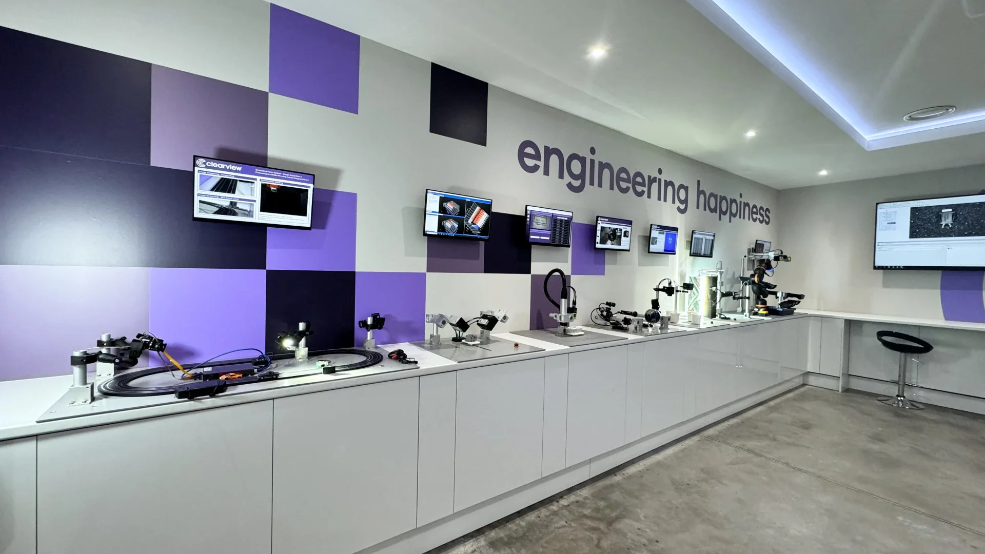 Modern engineering lab with microscopes, robotic arms, and computer monitors on a white counter. The wall has a purple and black geometric pattern and the words 'engineering happiness' displayed.