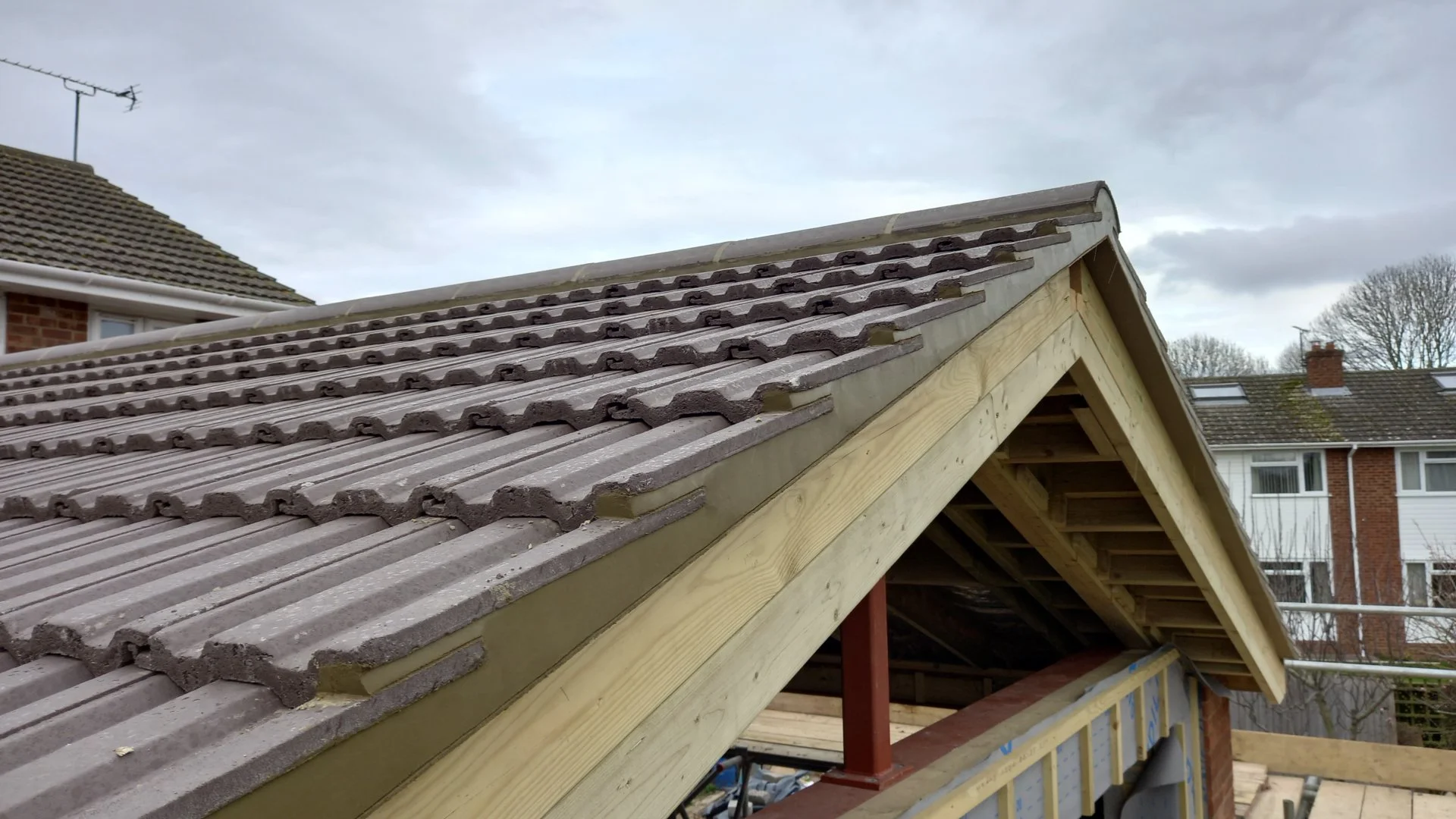 Partially constructed house roof with wooden framing and gray roofing tiles, under cloudy sky.
