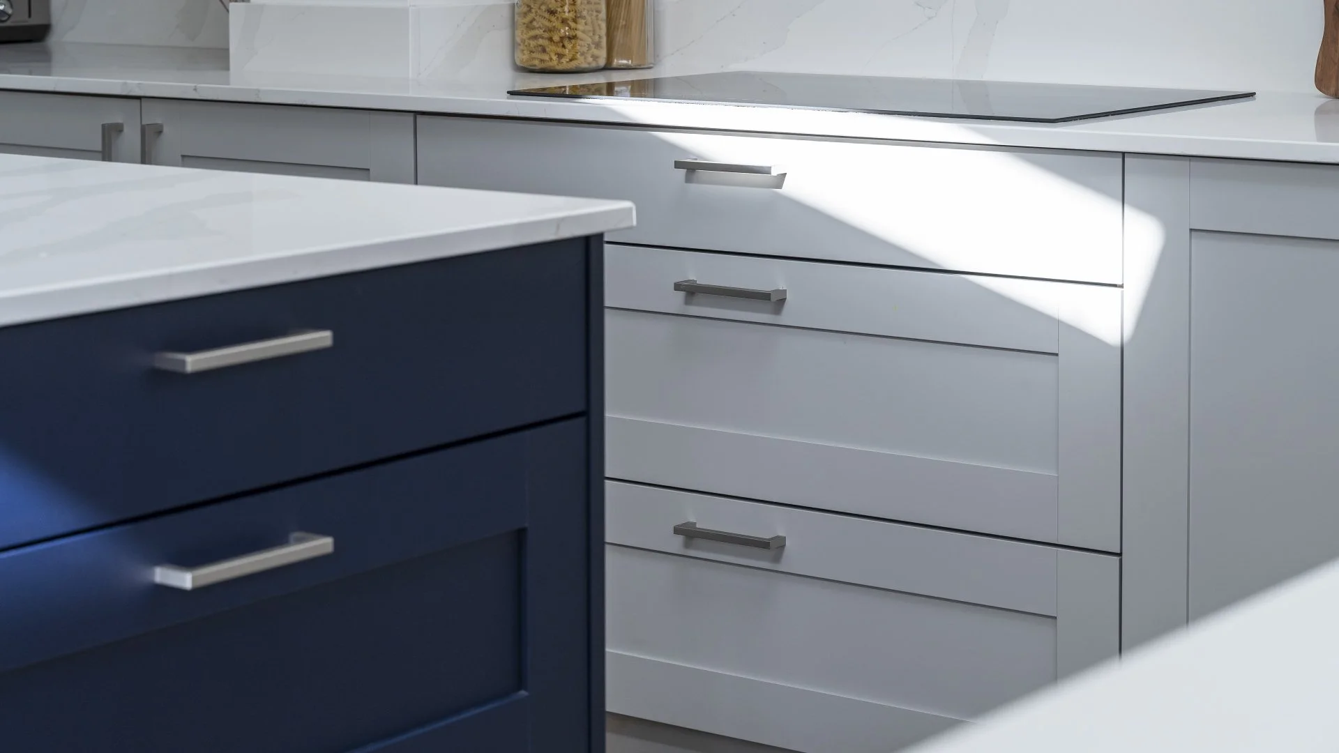 A modern kitchen with white cabinets, a blue island, and stainless steel handles, with sunlight casting shadows on the cabinets.