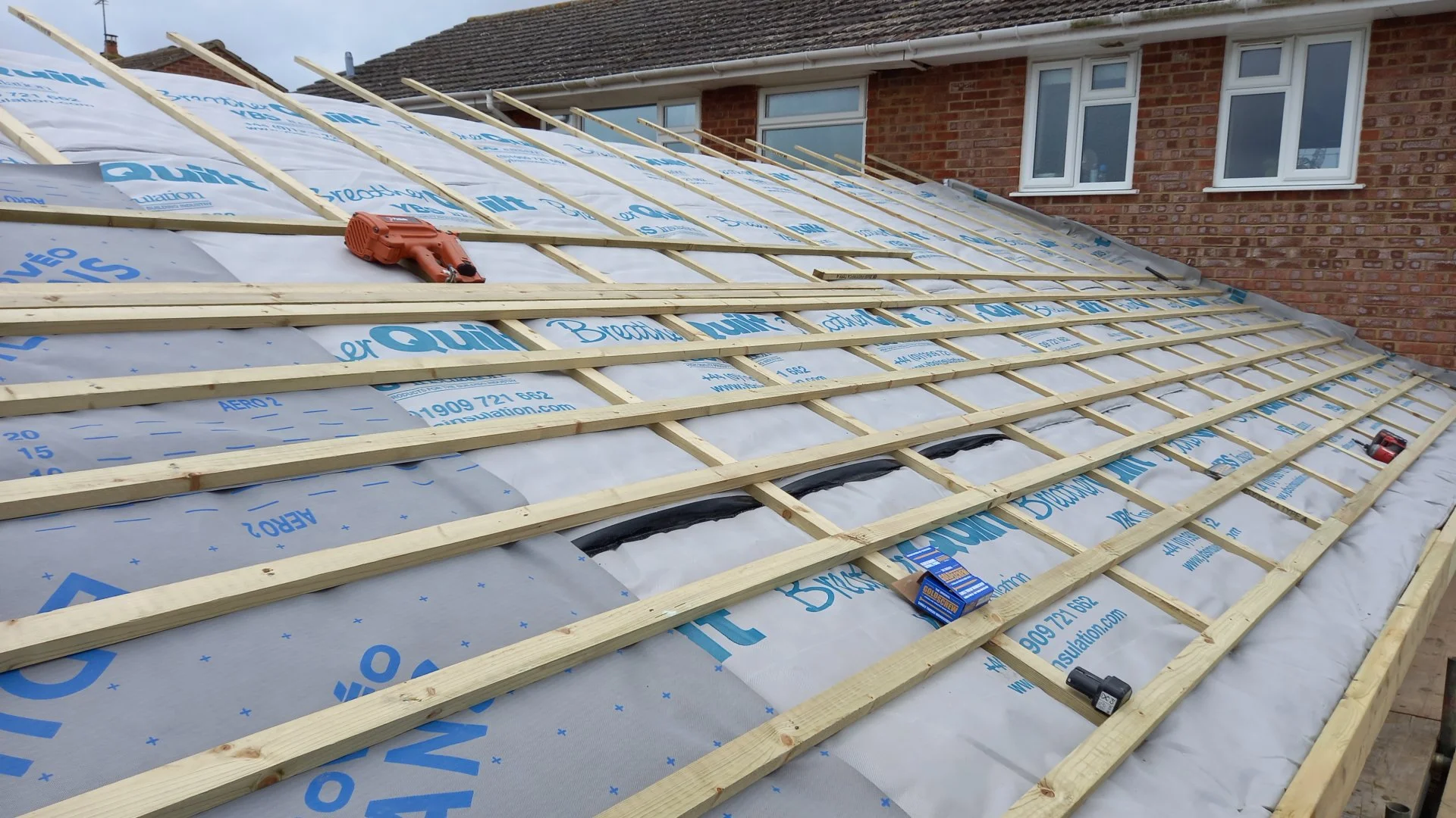Construction site showing a house roof with wooden battens installed for roofing, insulation material underneath, and some construction tools and supplies on the roof.