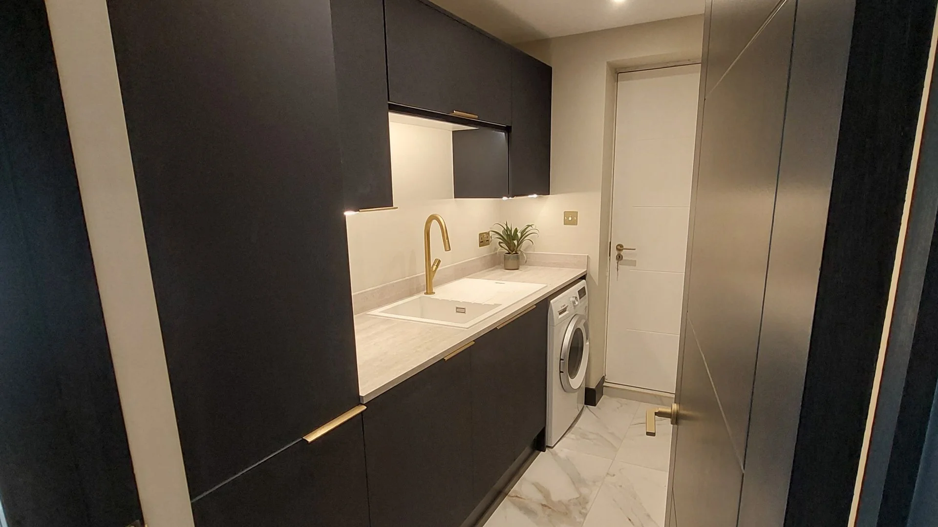 Modern laundry room with black cabinets, beige countertops, a gold faucet, a white sink, a potted plant, a washing machine, and a white door.