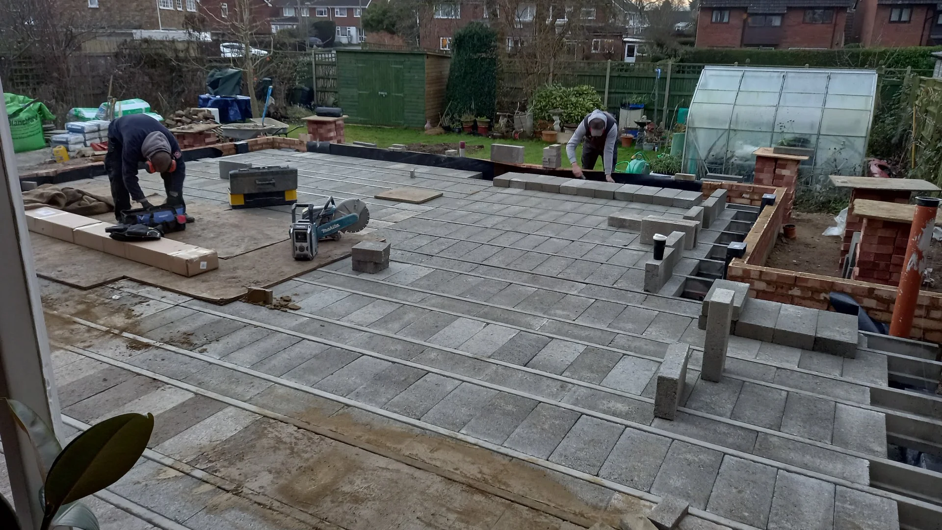 Two workers are laying concrete pavers on a backyard patio, with a partially constructed brick wall around the edge. Construction tools and materials are scattered around, including a saw, bags of cement, and pavers, with a green garden shed and gree