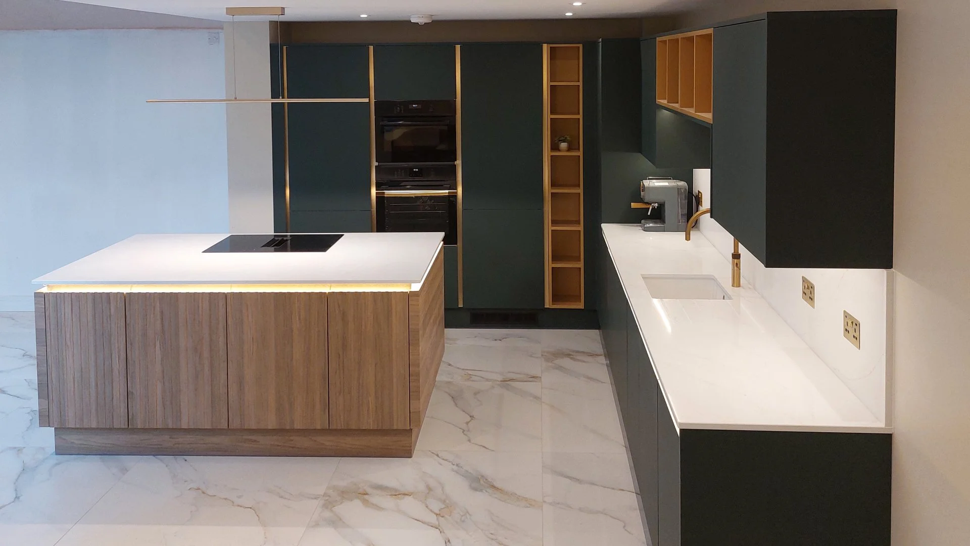 Modern kitchen with white countertops, dark green cabinets, and a wooden kitchen island with a built-in induction cooktop. There are built-in ovens, open shelving, a coffee machine, a sink, and marble flooring.