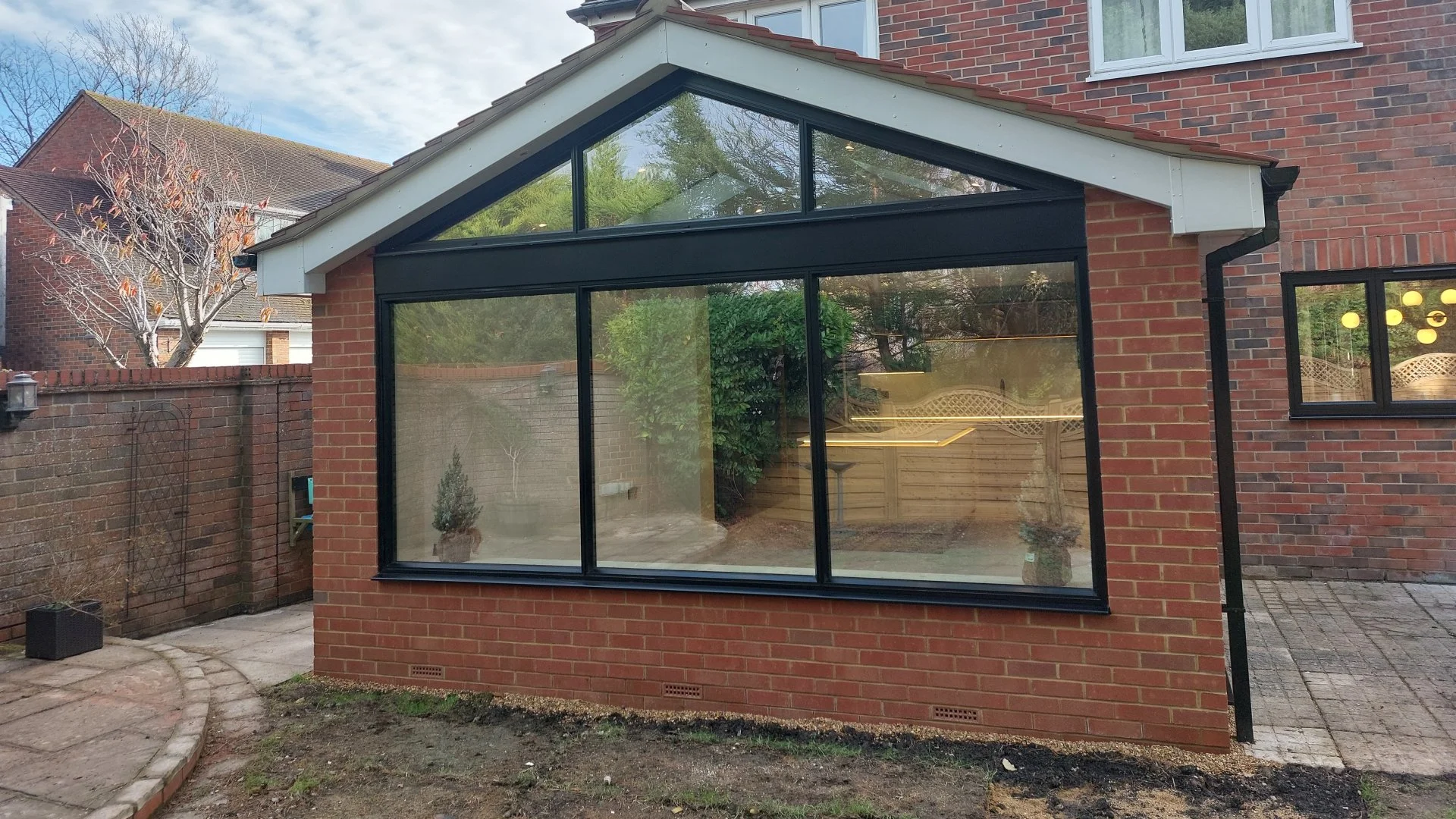Backyard with brick house featuring a large glass extension, a small paved patio, and a tree with sparse leaves.