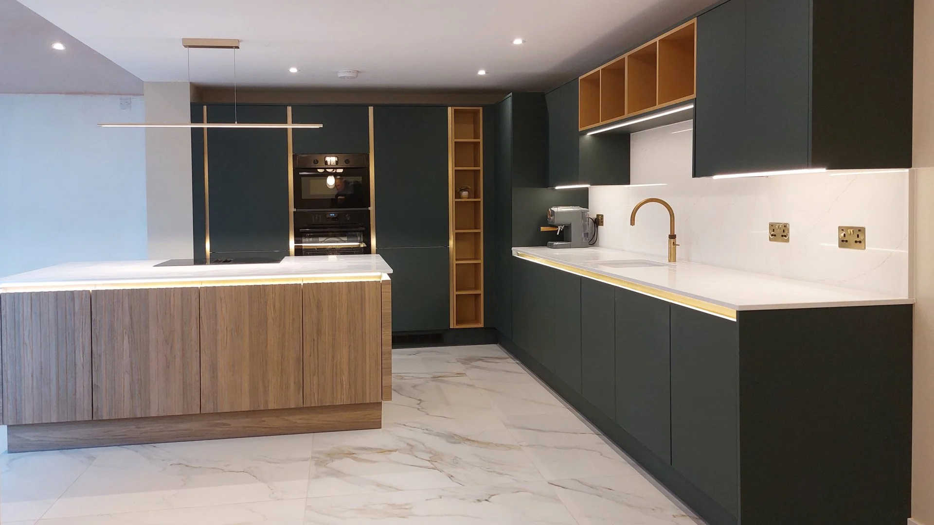 Modern kitchen with dark green cabinets, white countertop, gold accents, a built-in oven, coffee machine, and marble flooring.