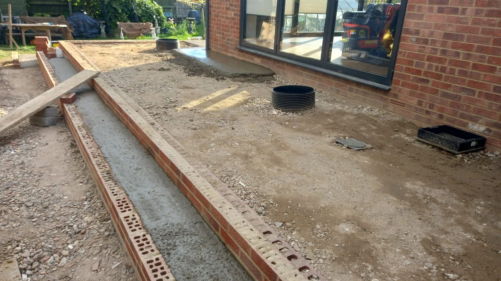 Construction site with brick edging and concrete work in progress next to a house with large windows and brick wall. There are construction materials and tools scattered around.