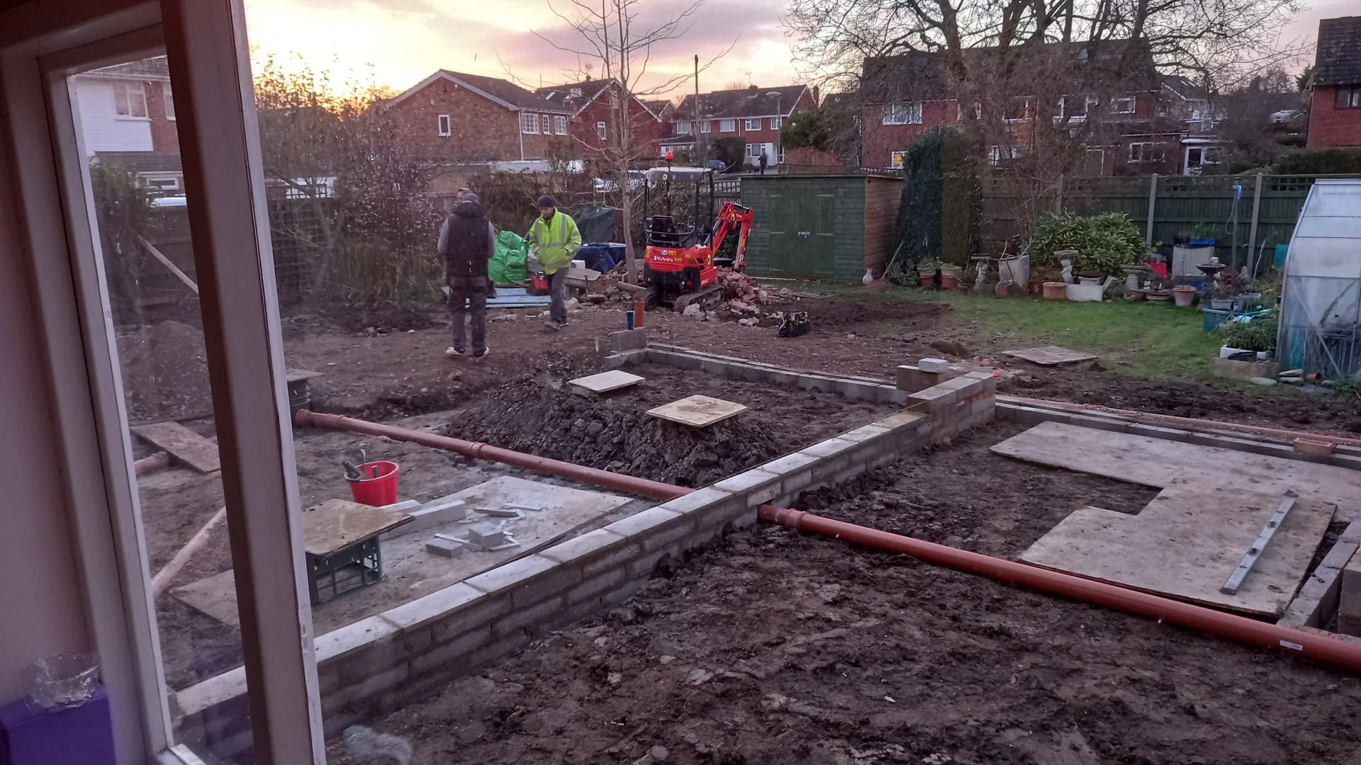 View of a backyard under construction, seen through a window. Two workers wearing high-visibility vests are working on the site, with construction materials, bricks, and a small excavator present. The yard is being prepared for future landscaping or 