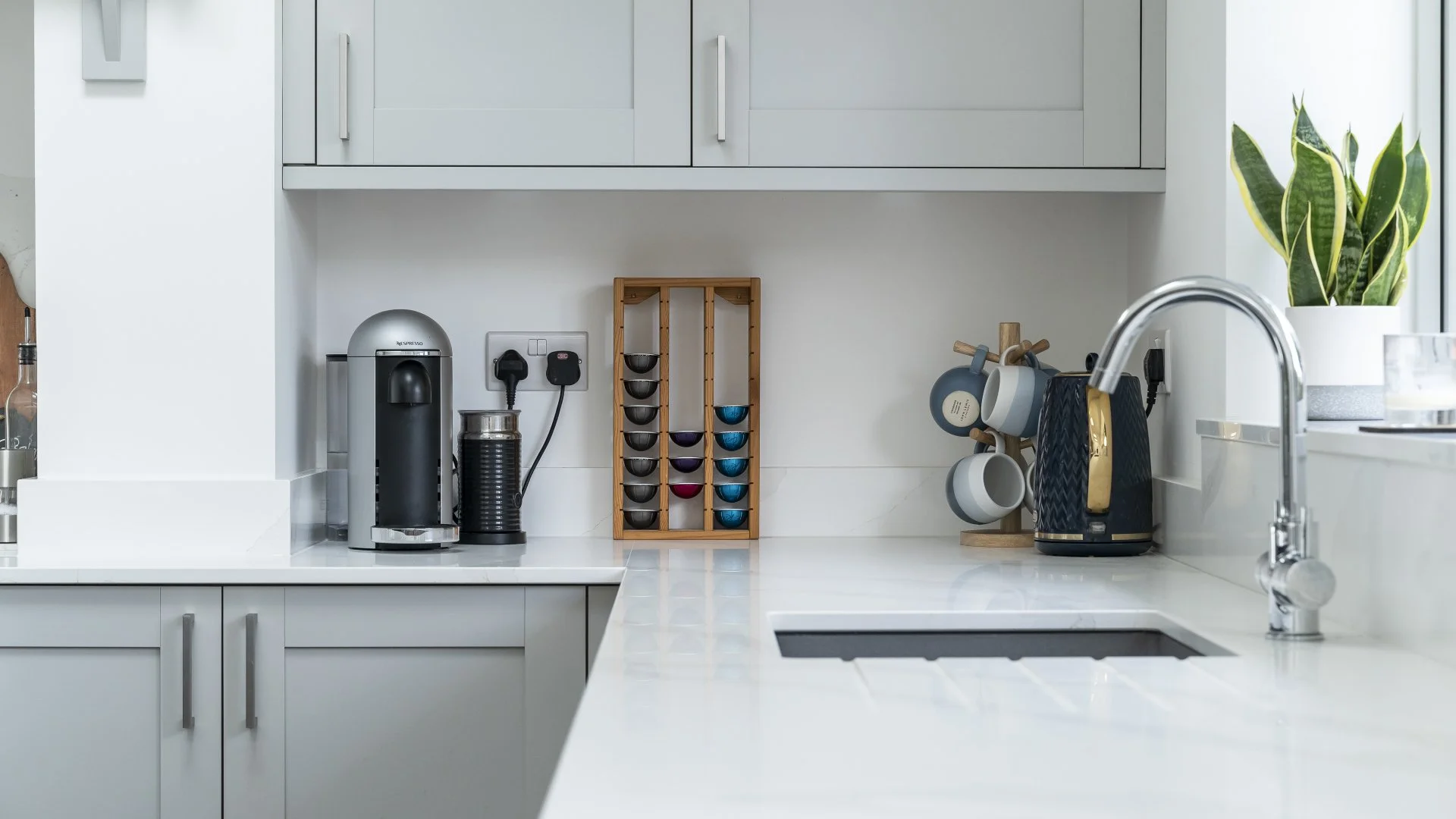 Modern kitchen with white countertops, grey cabinets, a coffee machine, a spice rack, and hanging mugs, with a plant near the window.