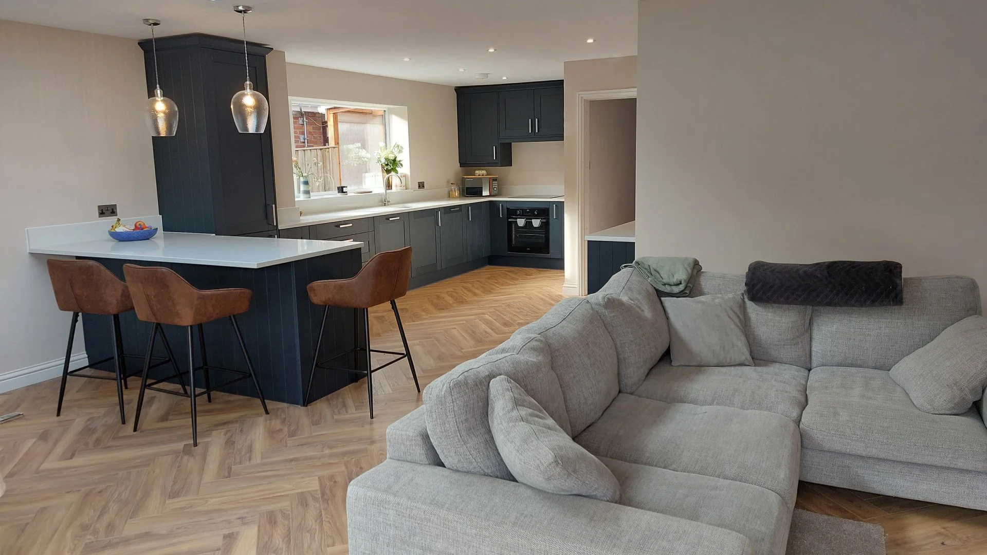 Open-plan living area with a kitchen and a cozy sofa. The kitchen features dark cabinets, a white countertop, a window with a plant, and three hanging pendant lights. The sofa is light gray with cushions and blankets, and there are three brown barsto