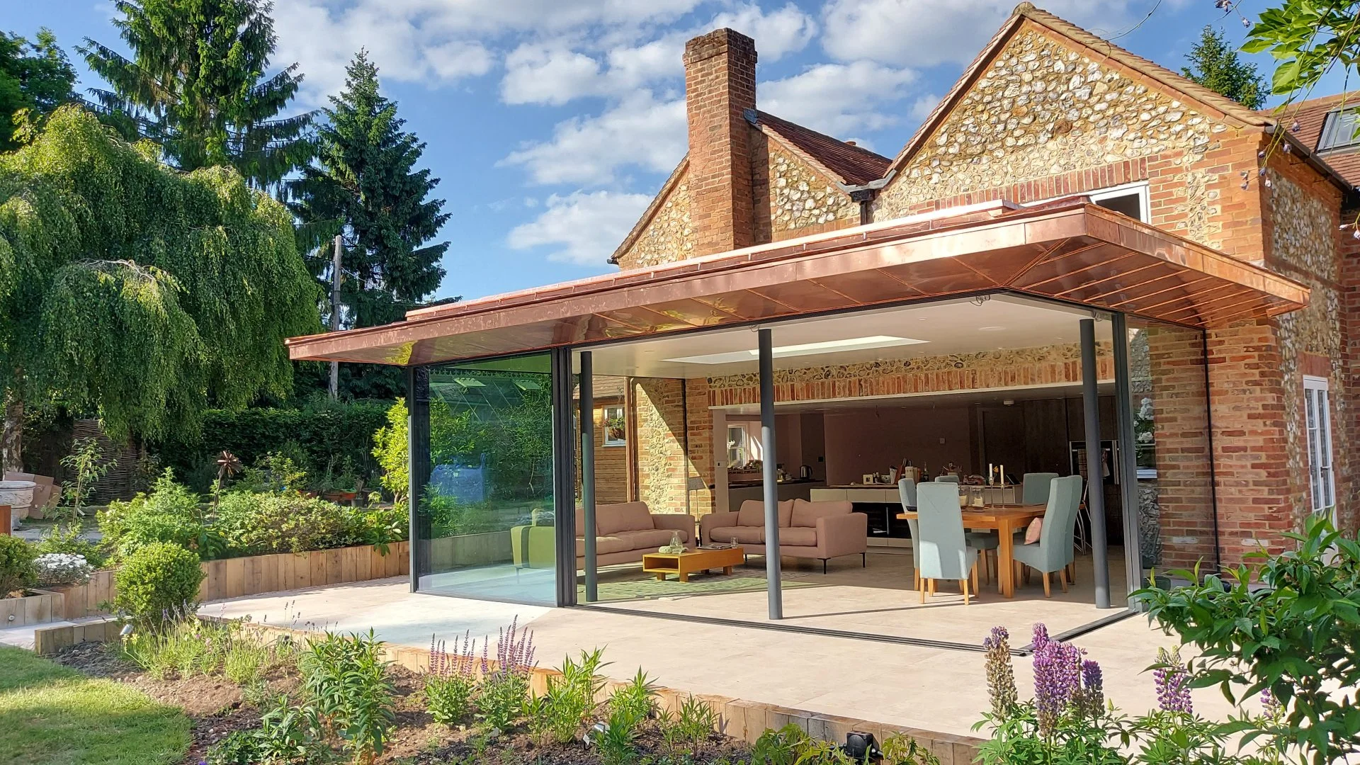 A modern backyard patio attached to a brick house with a glass enclosure, outdoor furniture, and lush greenery.