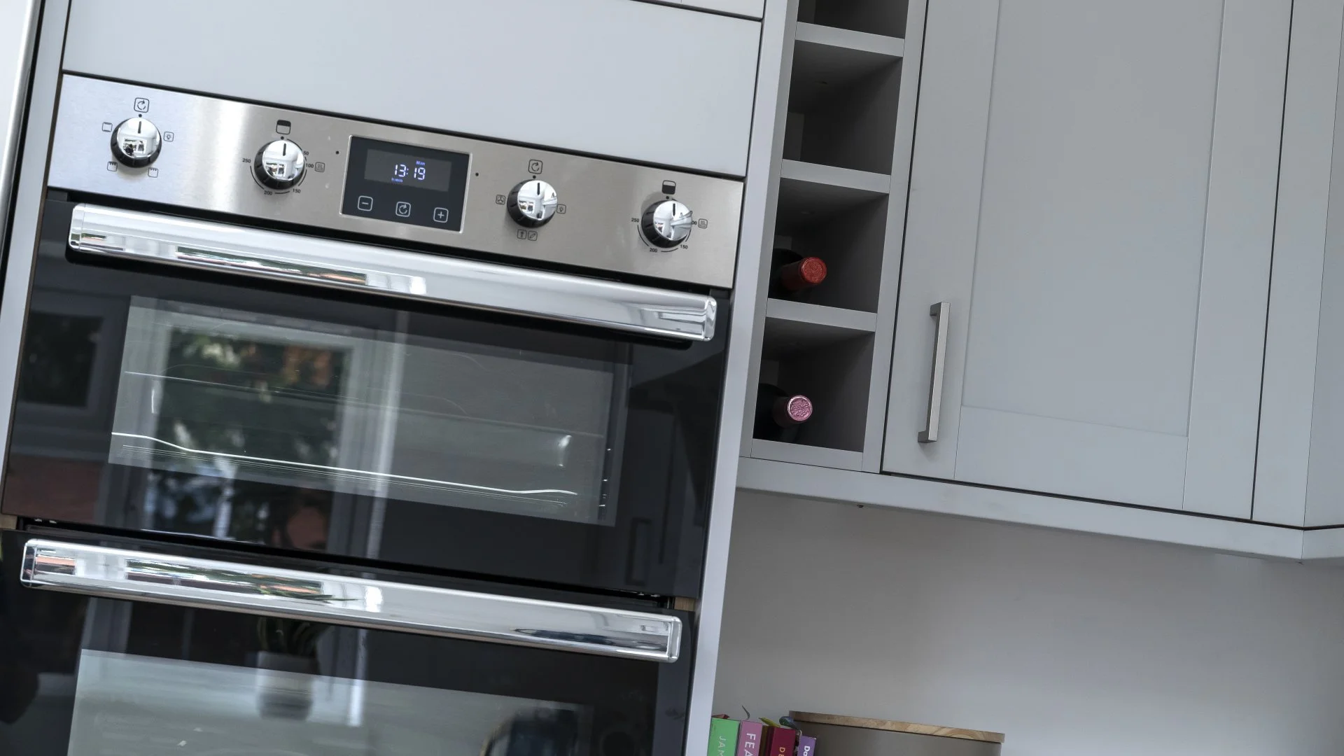 Modern kitchen with a stainless steel oven and a digital display, surrounded by white cabinets and open shelves with wine bottles.