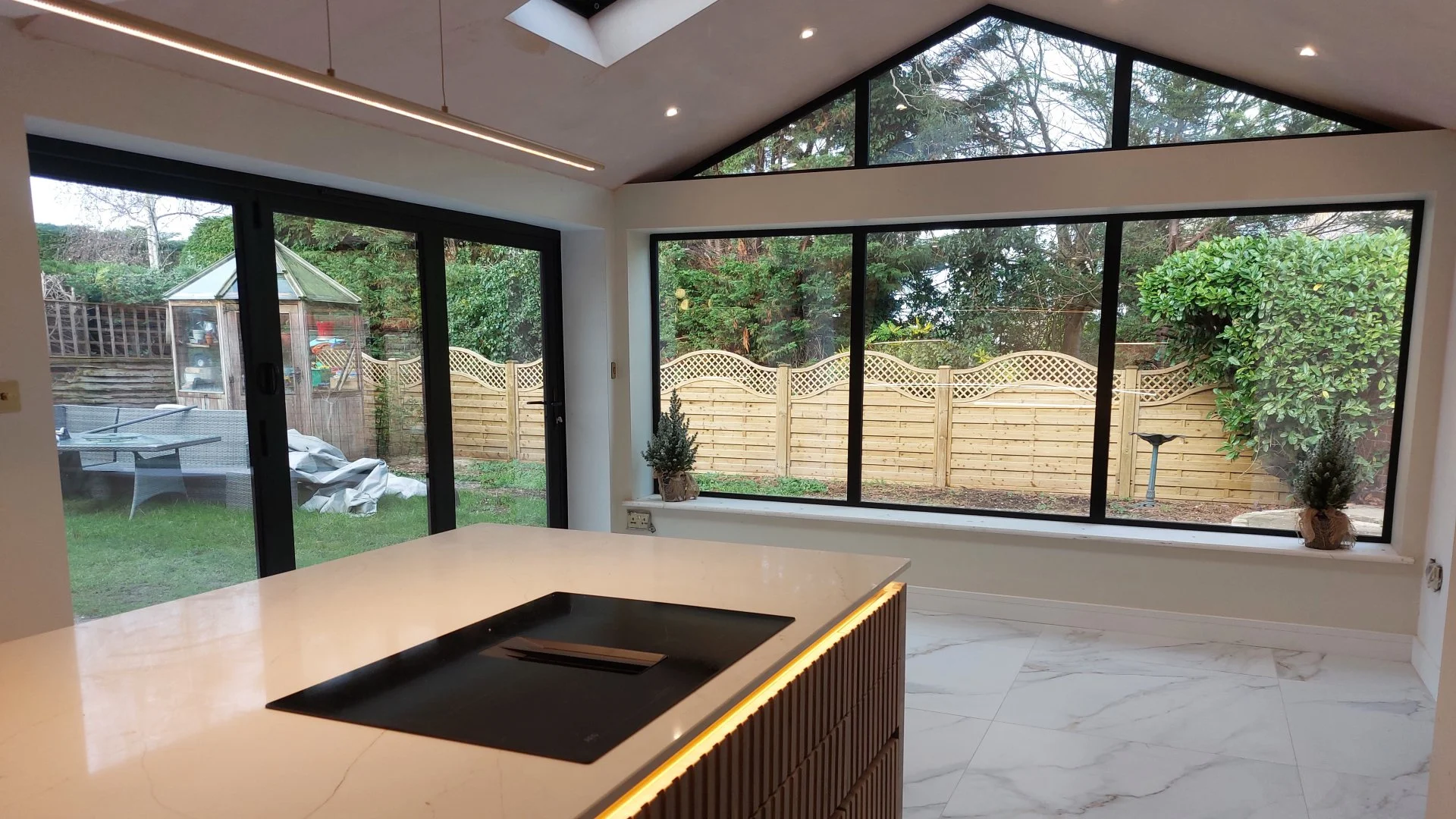 Modern kitchen with large floor-to-ceiling windows looking out onto a fenced backyard with trees and outdoor furniture. The kitchen features a white countertop with an integrated black cooktop and wooden lower cabinets, along with decorative plants o