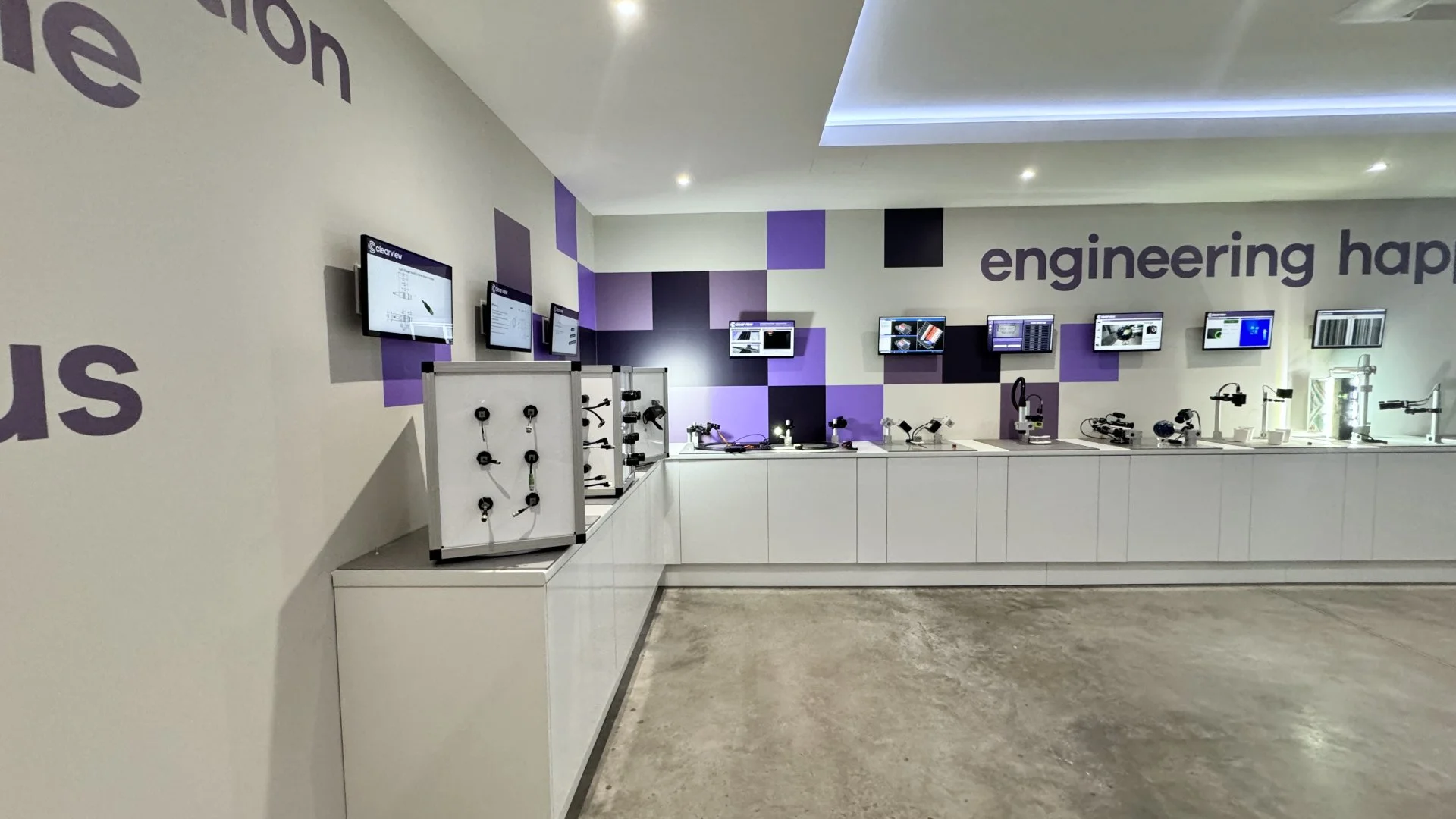 Modern exhibit of engineering and science equipment on white countertops, with monitors displaying technical diagrams and models on a purple and white wall.