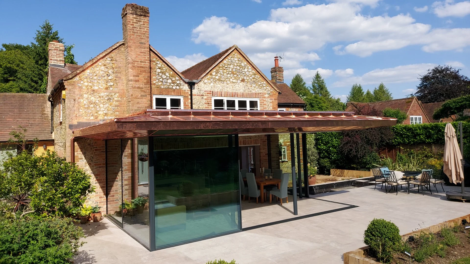 Backyard patio area with a modern glass enclosed room, outdoor dining table with chairs, sun umbrella, and well-maintained garden. Traditional brick house with pitched roof and chimney in the background, surrounded by lush greenery and a partly cloud