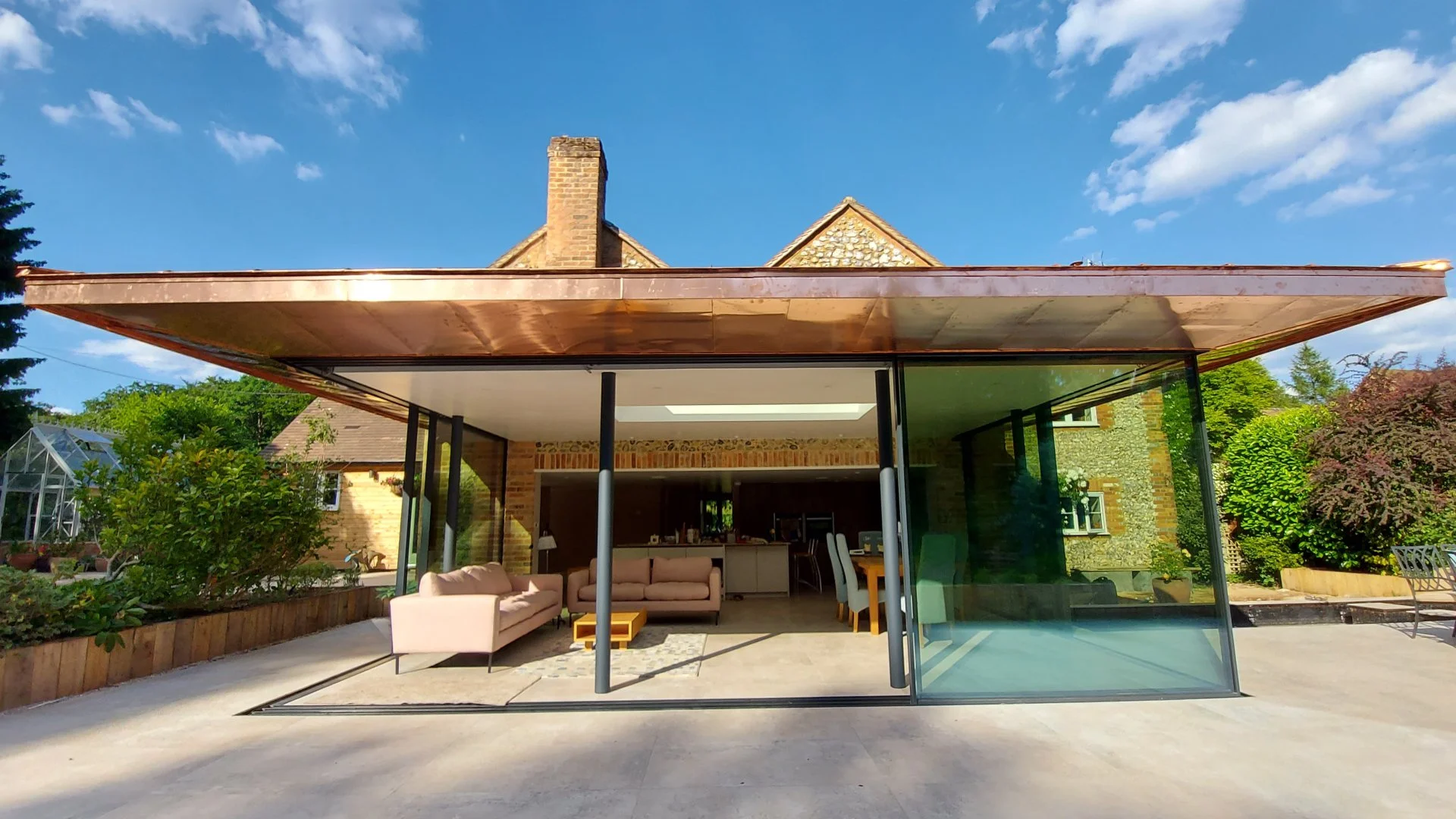 Modern house with an outdoor living area featuring a beige sofa and chairs, glass sliding doors, brick and stone exterior, surrounded by greenery and a blue sky.