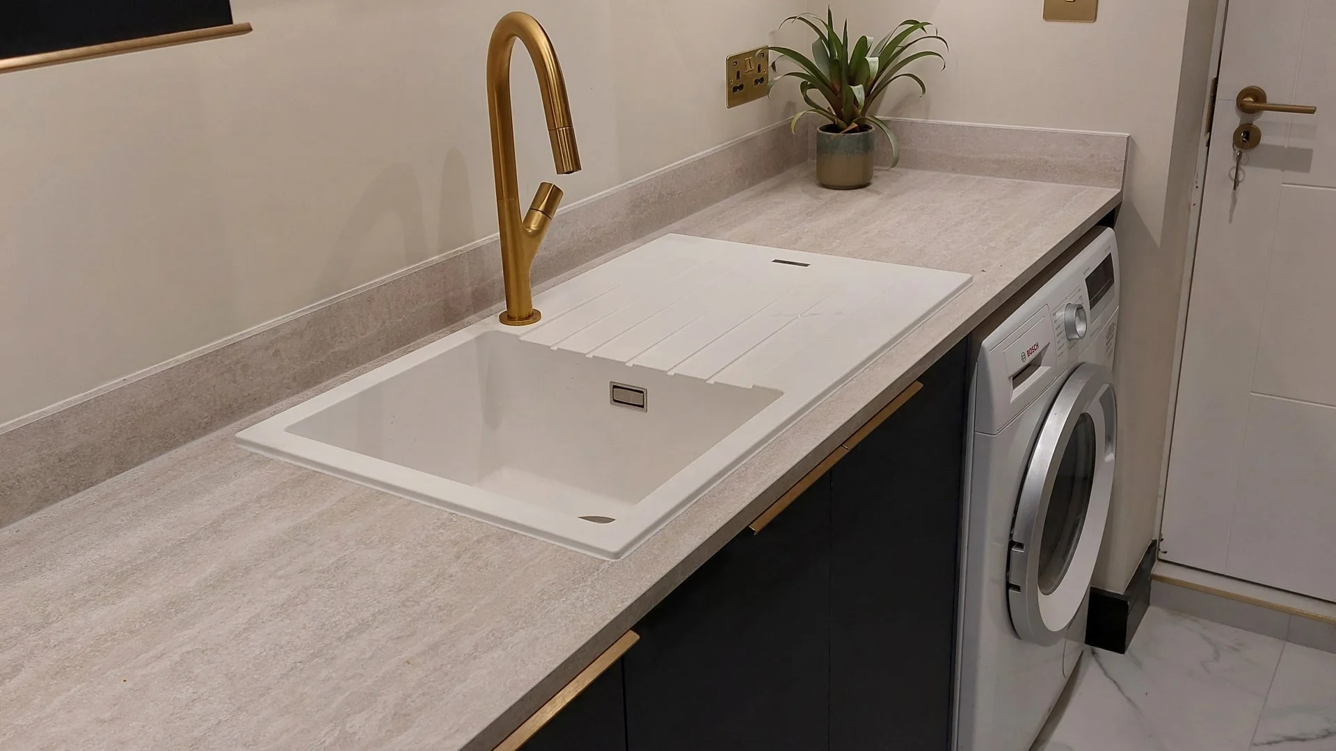 Kitchen laundry area with beige countertop, white sink, copper faucet, potted plant, washing machine, and utility cabinet.