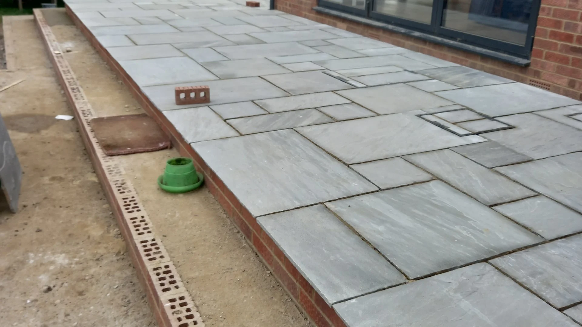 Newly laid gray stone patio with brick border next to a brick house with large windows. Construction materials and tools are present, including a level, brick, green pipe, and drainage grate.
