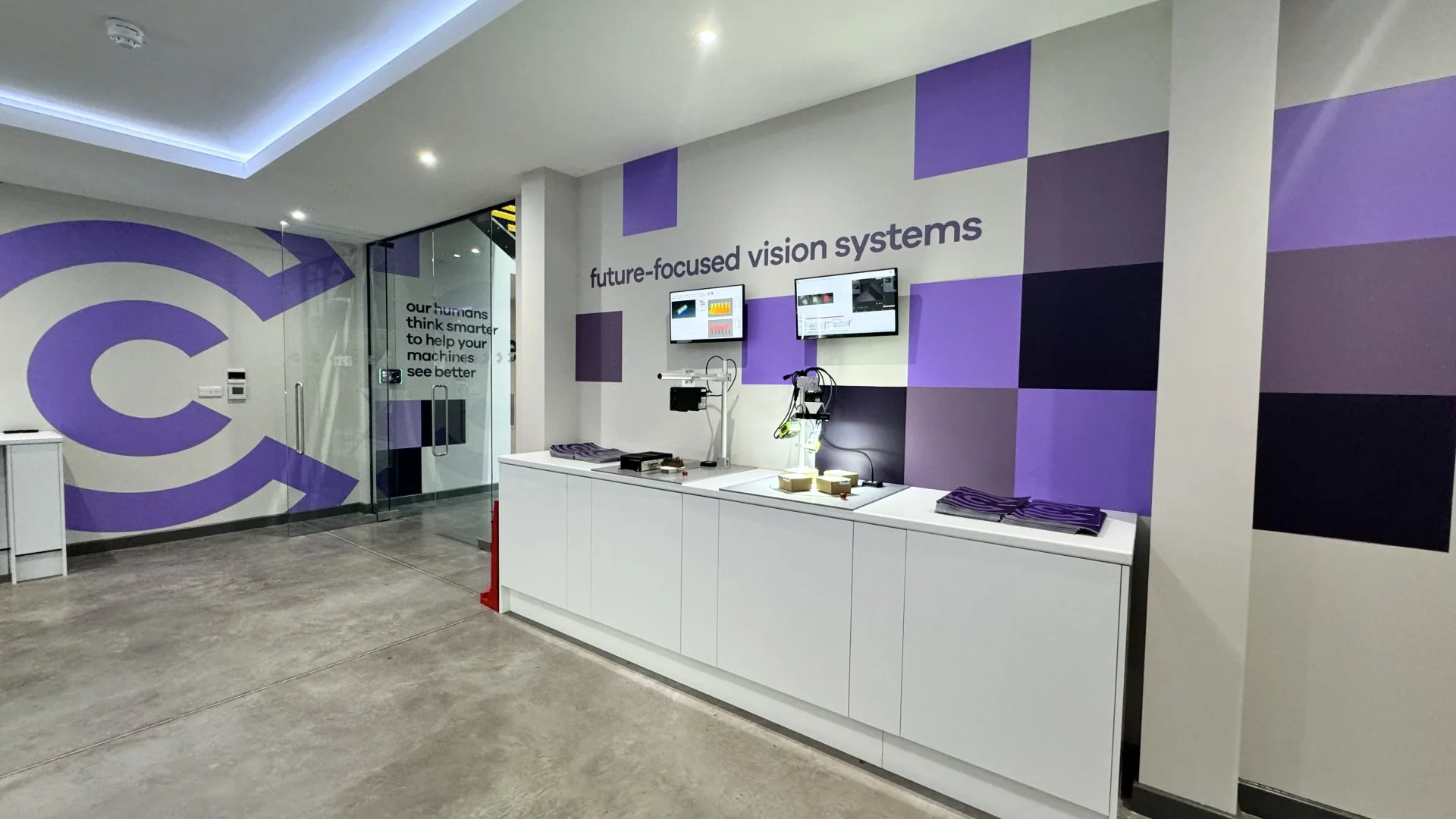 Modern office or showroom with a purple and white theme, featuring a wall with the text 'future-focused vision systems', two monitors, and a glass door with the words 'our humans think smarter to help your machines see better'.