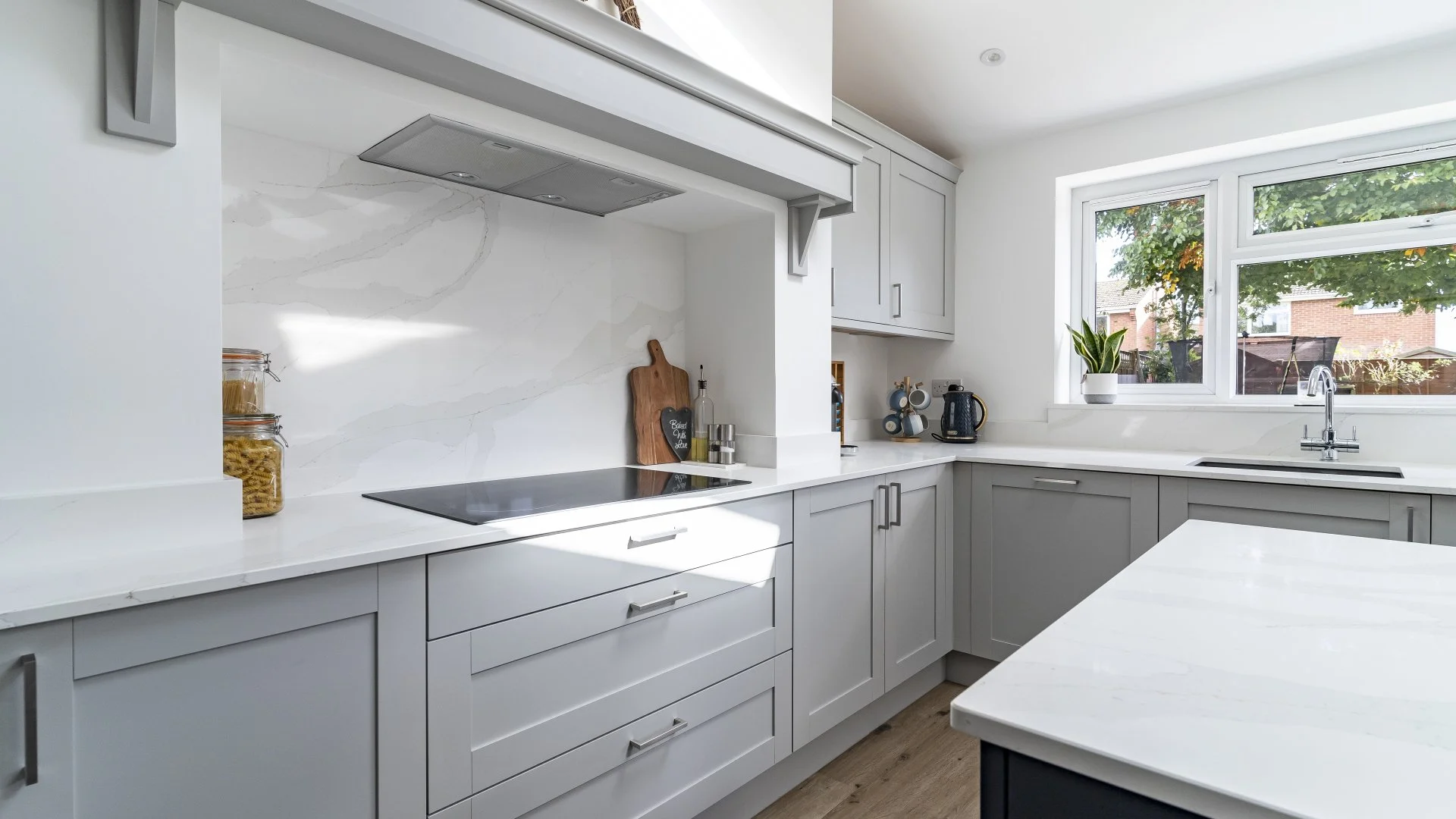 A modern kitchen with white countertops, light gray cabinets, a window overlooking a garden, a black kettle, a potted plant, jars of pasta, and a cutting board on the counter.