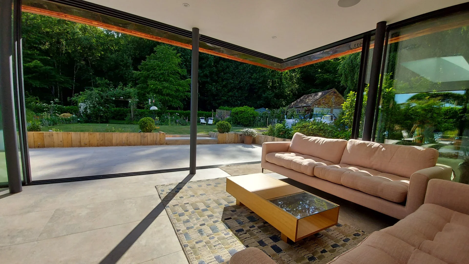 View of a backyard garden seen from a living room with large glass doors. The garden features a lush green lawn, trees, outdoor furniture, and a small shed or garage in the background.