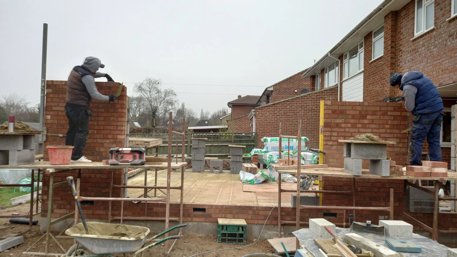 Two construction workers building a brick wall outside of residential houses on a cloudy day, with construction tools and materials scattered around.