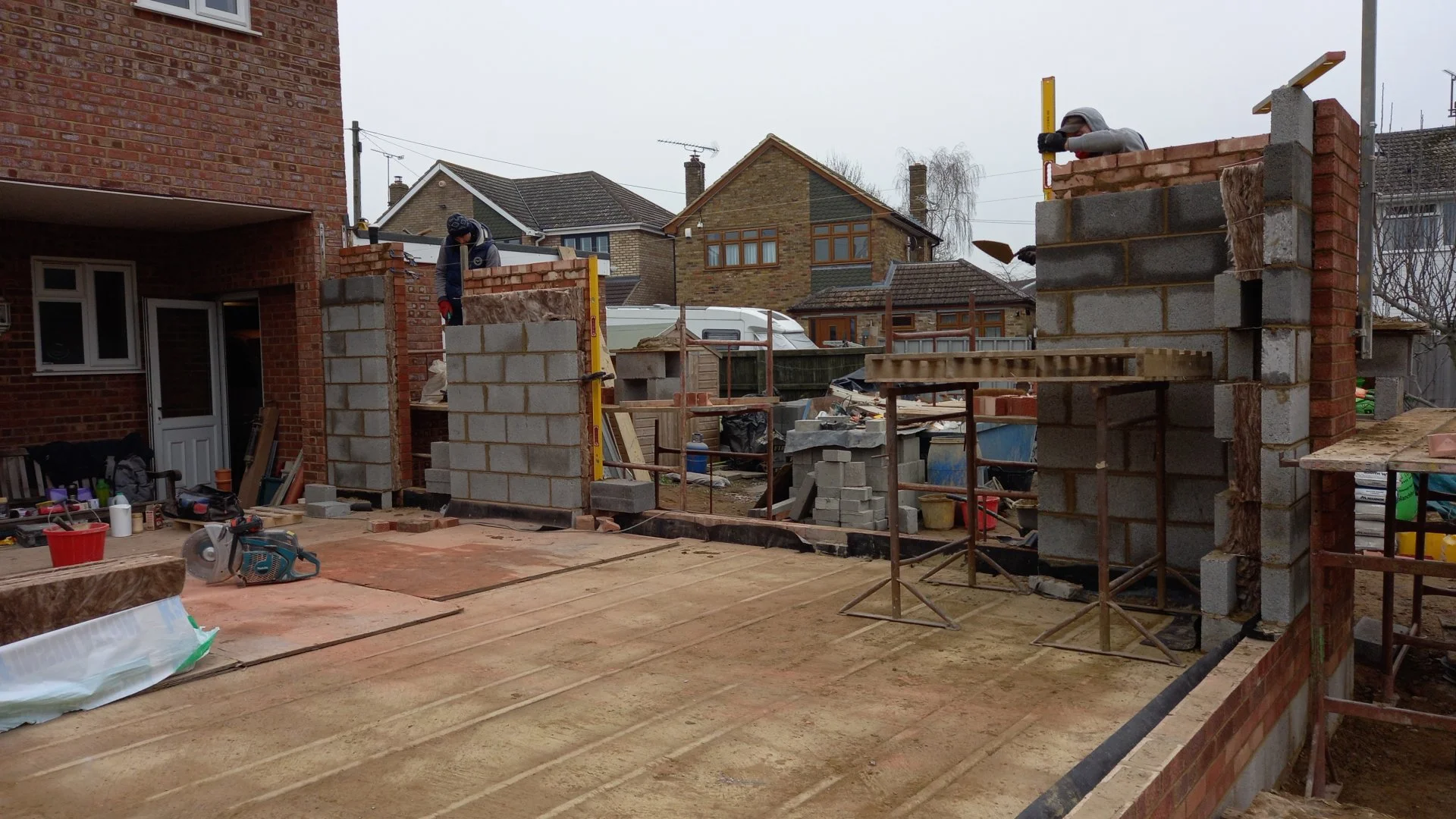 Construction workers building brick wall on residential construction site. Tools and materials scattered around, with existing houses in the background.