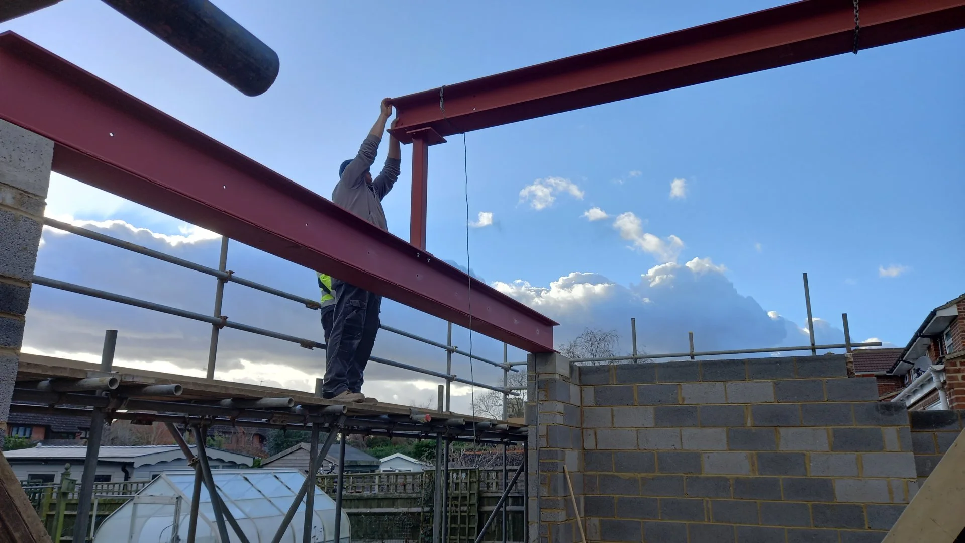 Two construction workers are installing a steel beam on a building under construction. One worker is standing on scaffolding, and the other is holding the beam in place. The framework of the building is visible, with a brick wall in the foreground an
