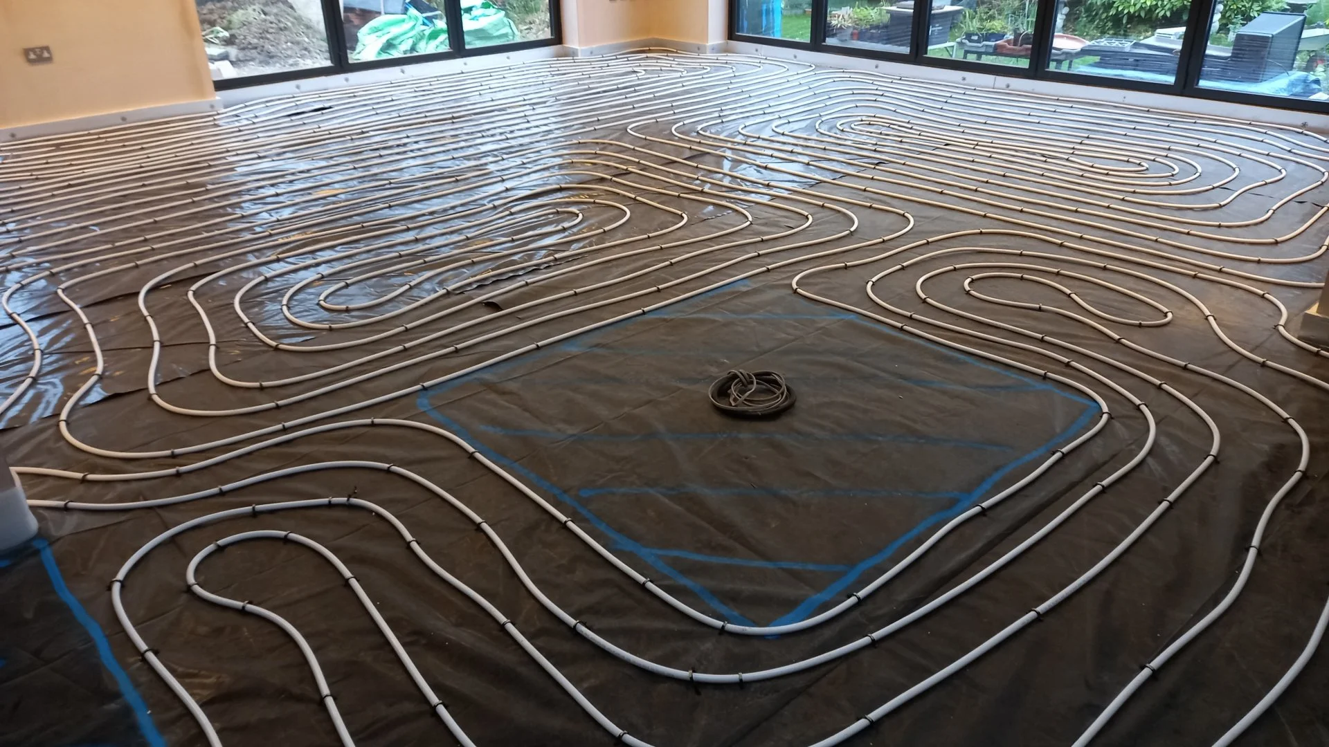 Underfloor heating pipes installed on a dark plastic vapor barrier, with large windows showing an outdoor garden space.
