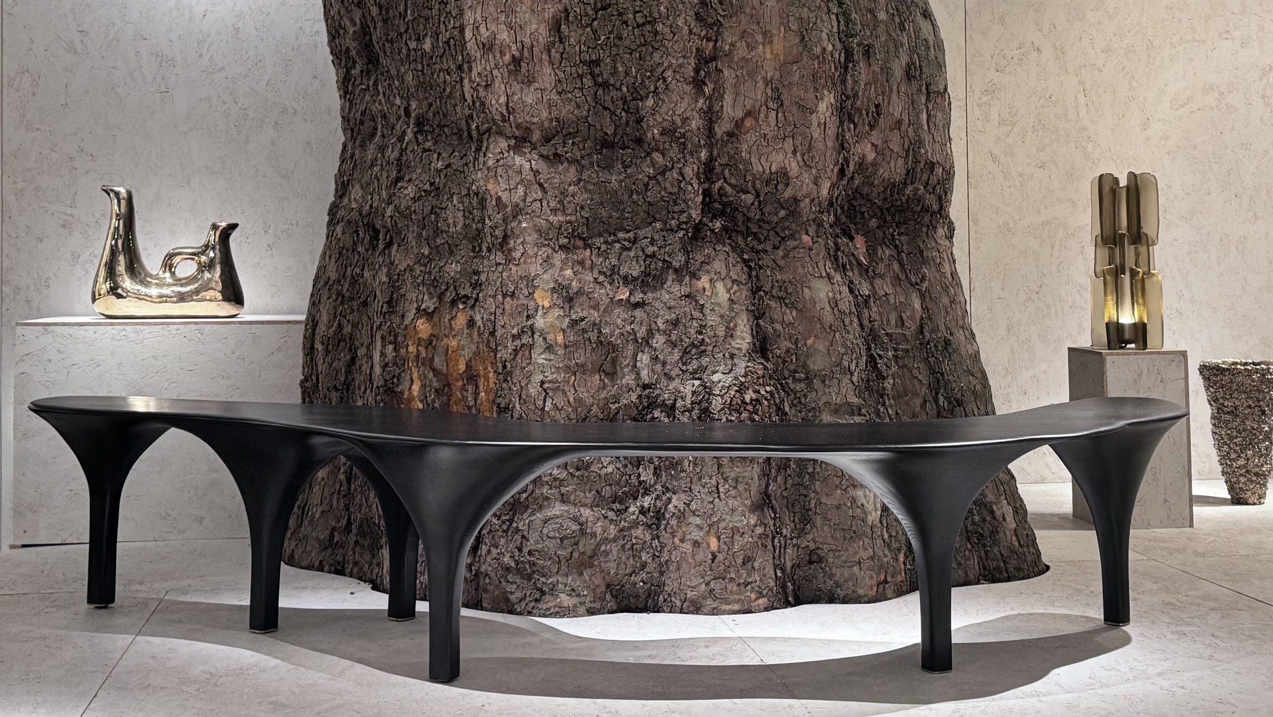 Un banc noir moderne avec des supports sinueux placé devant un arbre grand en intérieur au PAD London 2025. À l'arrière, des sculptures décoratives dorées et une grande urne en pierre décorative.