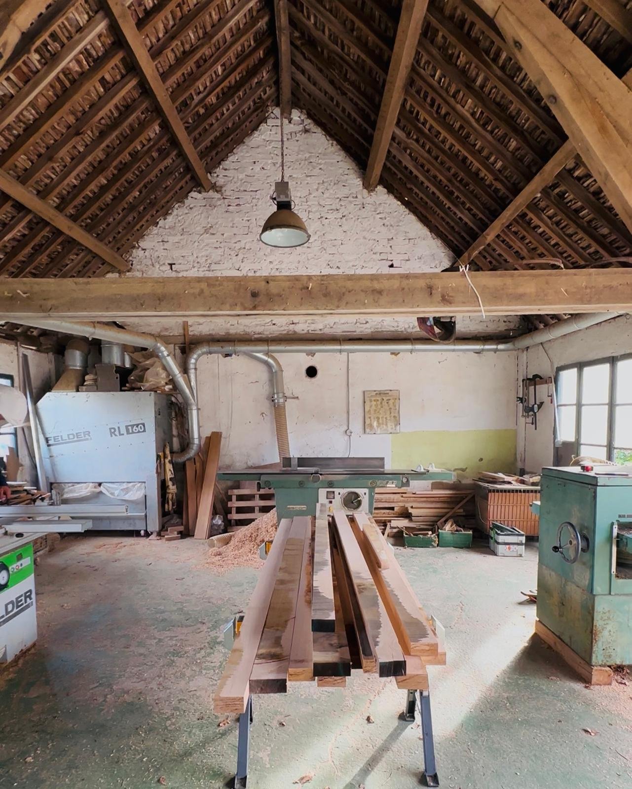 Un atelier de menuiserie avec une table de travail et des planches de bois, entouré d'outils, machines et matériaux de bois, dans un espace lumineux avec fenêtres et un plafond en pente en bois.