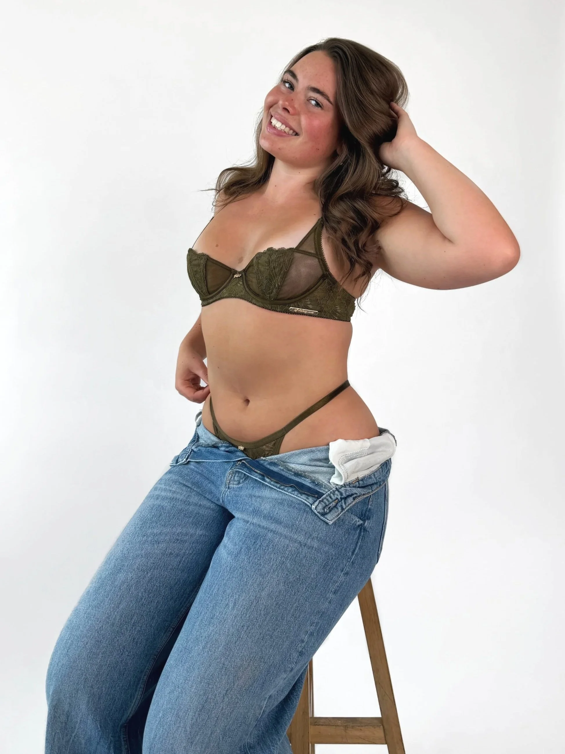 A woman with long brown hair smiling, sitting on a wooden stool, wearing blue jeans and an olive green lace bra, against a plain white background.