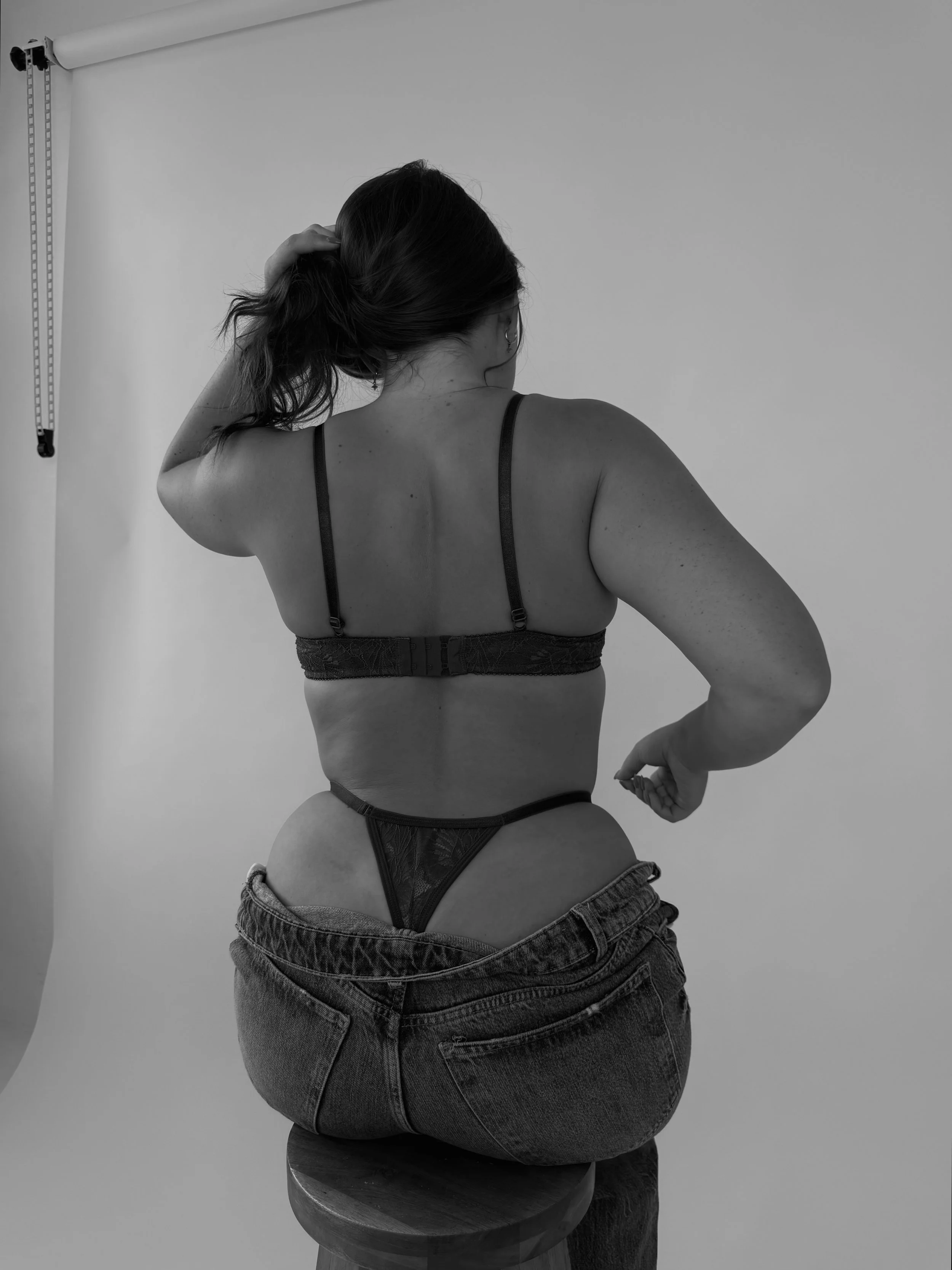 Black and white photo of a woman sitting on a round wooden stool, seen from the back, with her right arm raised and her hand touching her hair. She is wearing a bra and high-waisted jeans, with her jeans slightly pulled down, exposing the waistband of her underwear.