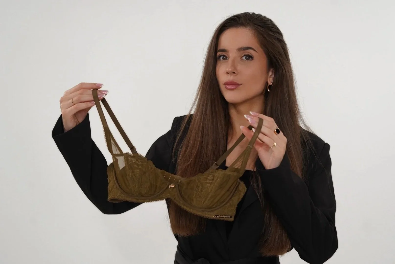 Woman with long brown hair, wearing a black blazer, holding a brown lace bra with both hands in front of a plain white background.
