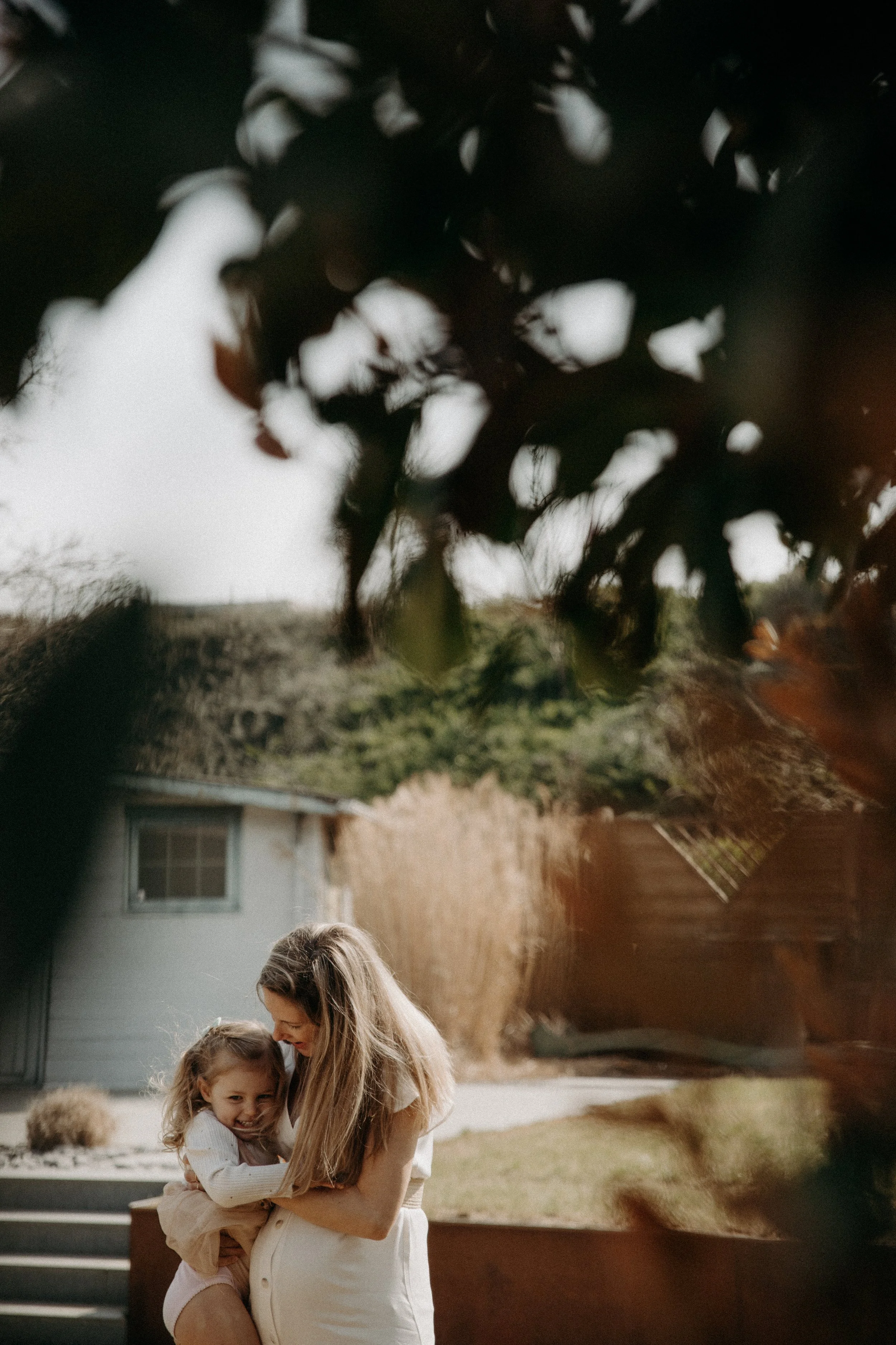 Une femme tient une fille dans ses bras, tous deux souriants, dans un jardin résidentiel, parmi des arbres et des buissons.