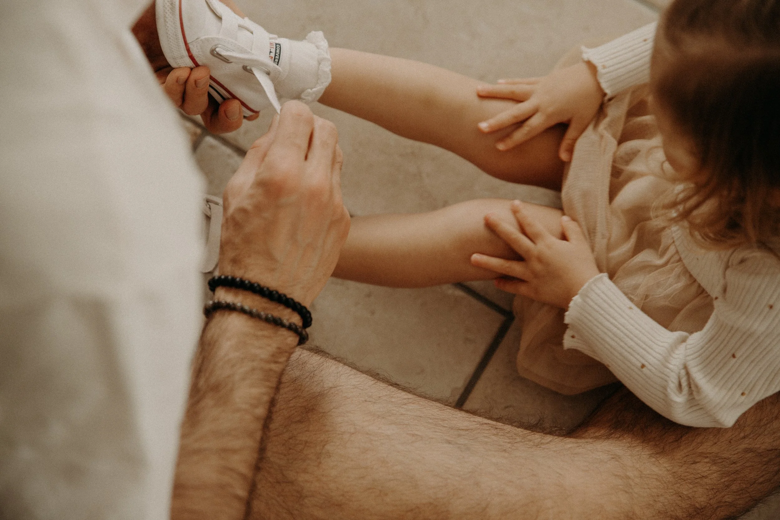 Une personne adulte attache les lacets d'une chaussure blanche à une enfant qui est assise par terre, vêtue d'une robe beige.